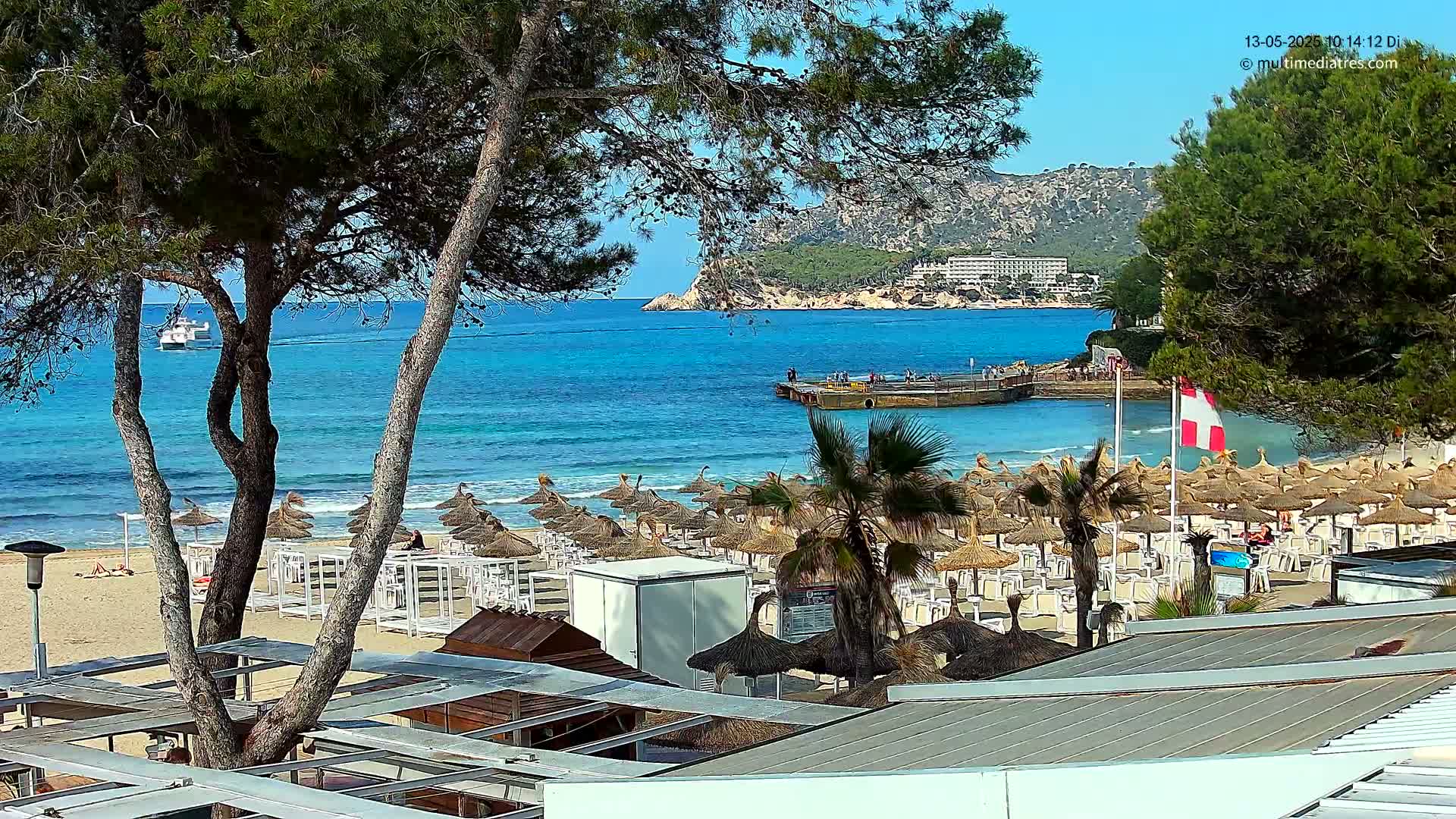 A sandy beach with numerous thatched umbrellas and lounge chairs is situated beside a calm, turquoise ocean under a clear, sunny sky, with a distant shoreline and a boat visible on the water.