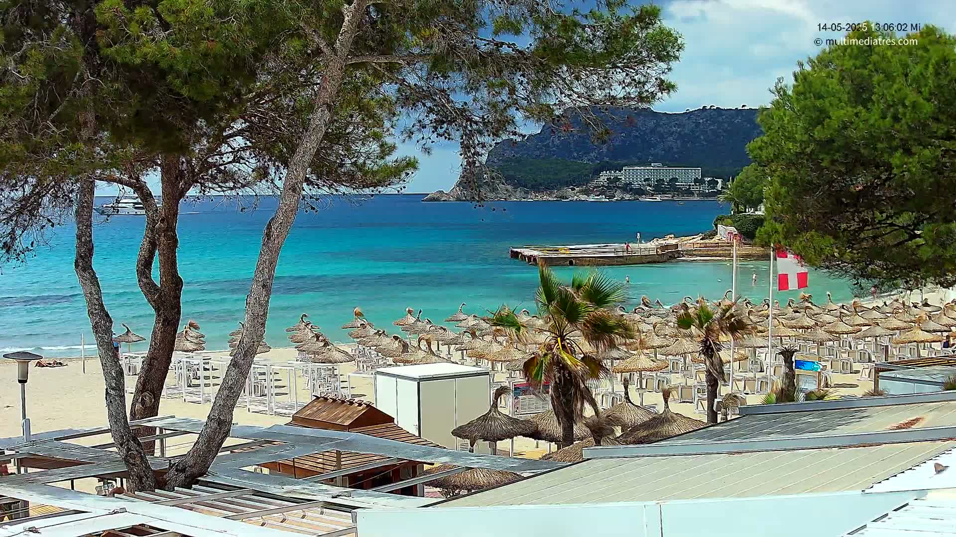 A sandy beach with numerous thatched umbrellas and palm trees overlooks a calm, turquoise ocean and a distant rocky hillside under a partly sunny sky.