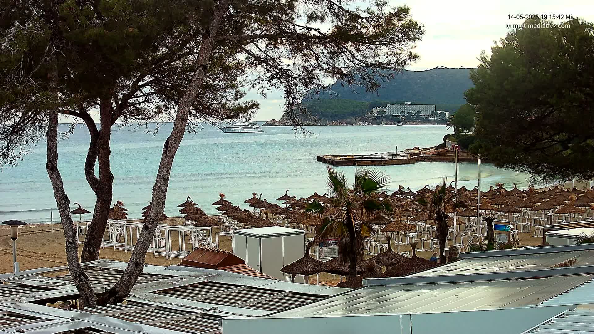 A sandy beach with numerous thatched umbrellas and beach chairs sits next to calm, light-blue water under a partly cloudy sky, with a boat and distant buildings visible in the background.
