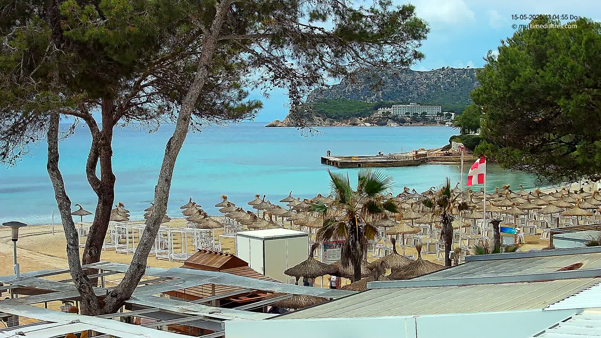 A sandy beach with numerous thatched umbrellas and white lounge chairs overlooks a calm, turquoise ocean under a partly cloudy sky, with a distant hotel and green hills visible.