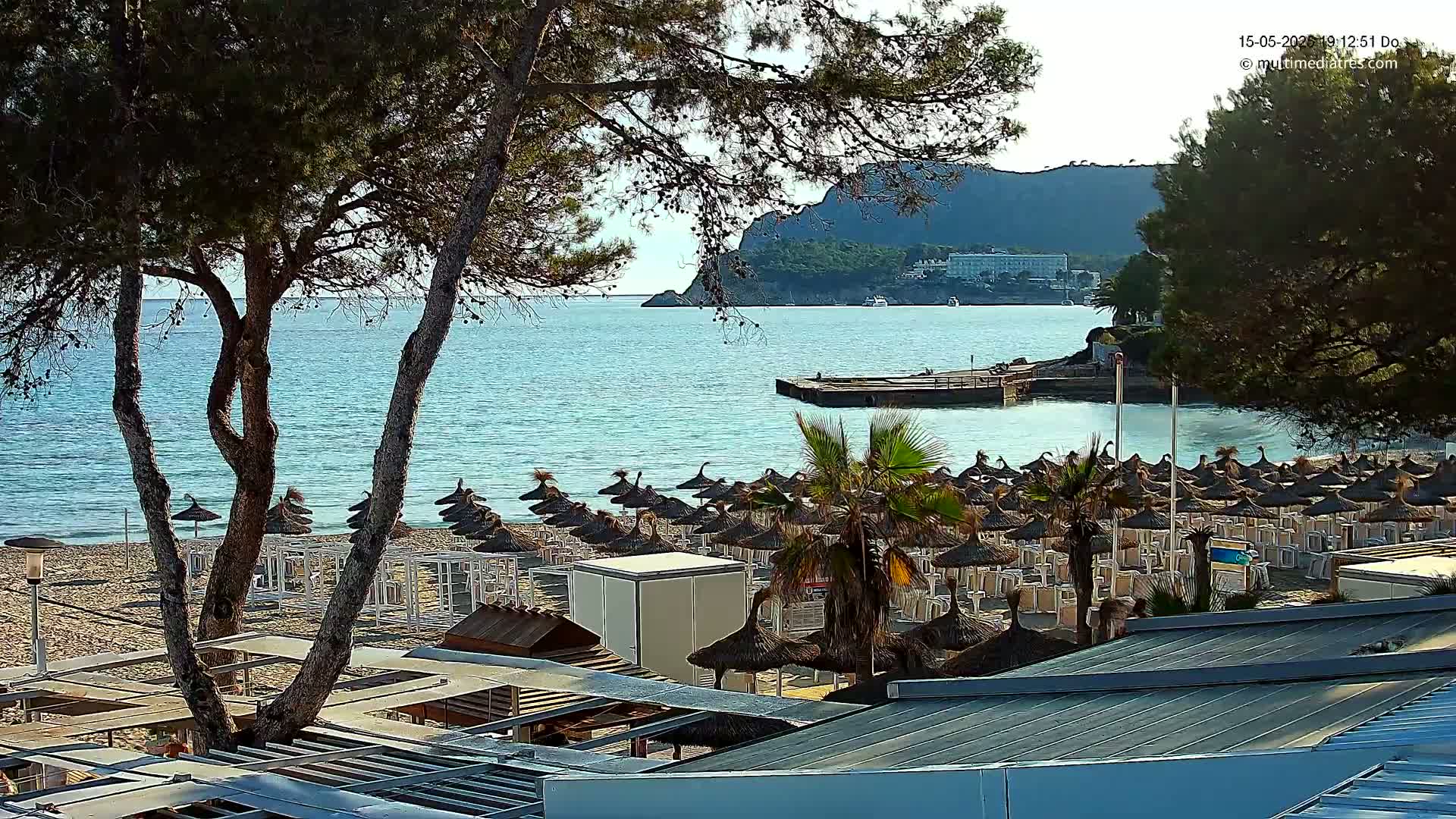A sandy beach with numerous thatched umbrellas and lounge chairs is situated beside a calm, blue sea, overlooked by a rocky hillside under a clear sky.