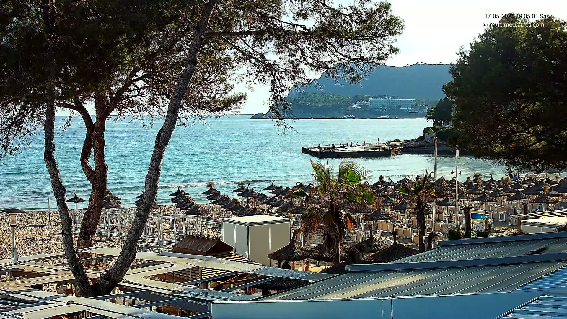 A sandy beach with numerous thatched umbrellas and lounge chairs is situated next to a calm, turquoise ocean, backed by a hilly coastline under a clear, sunny sky.