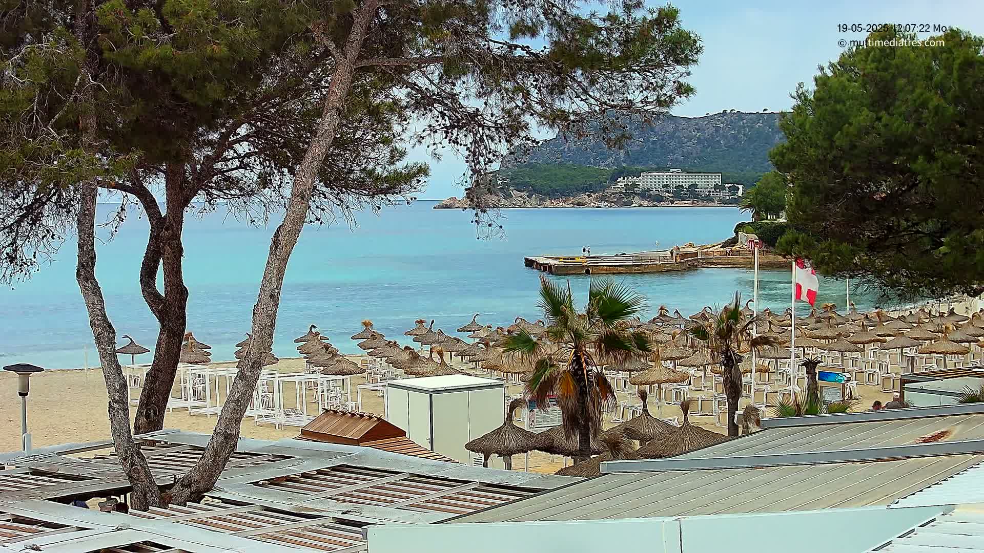 A sandy beach with numerous thatched umbrellas and lounge chairs is situated next to a calm, turquoise sea under a mostly sunny sky, with a tree partially obscuring the view.