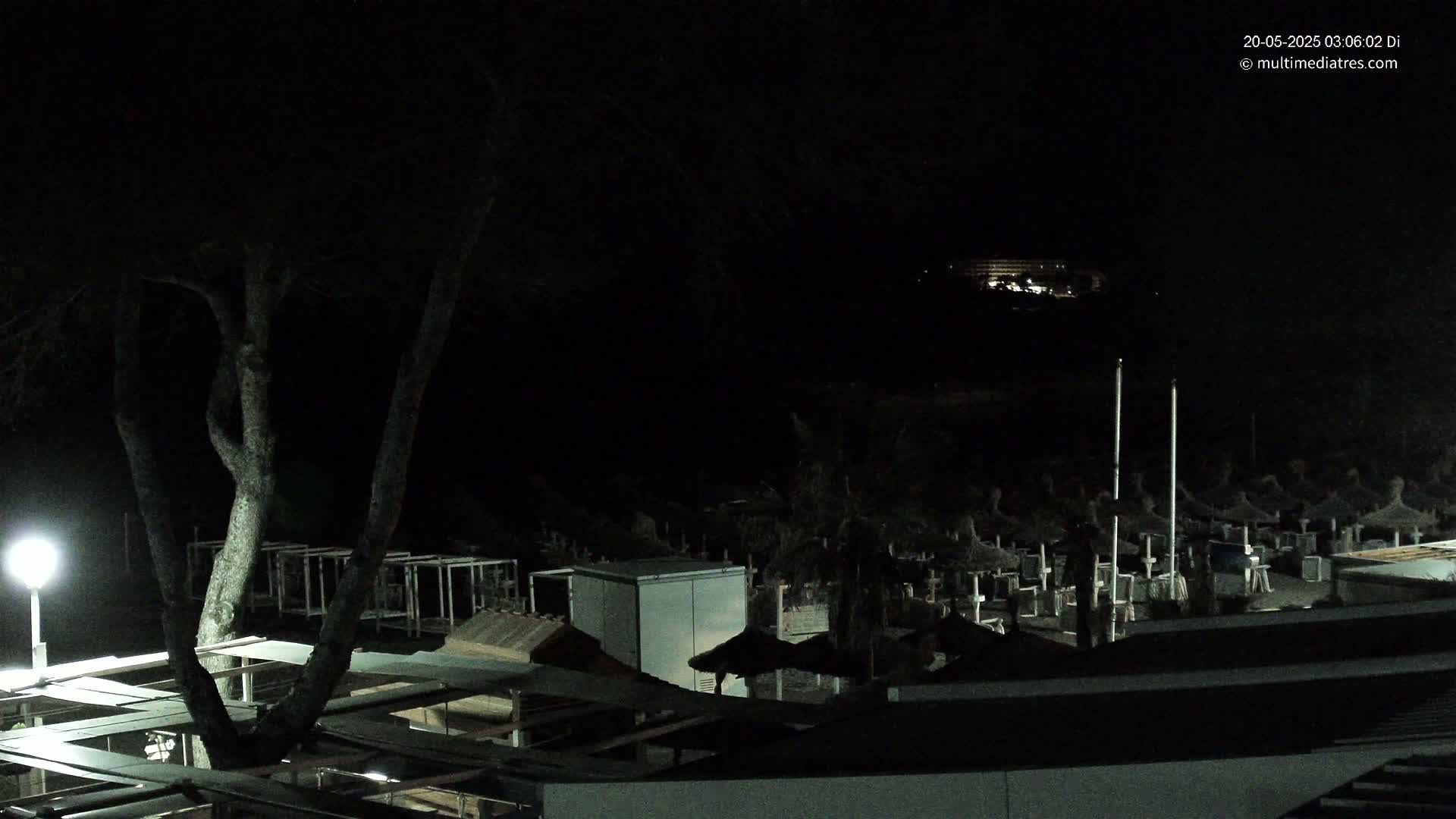 A nighttime view of a beach at night, showing closed beach umbrellas and a large building illuminated in the distance under a dark sky.