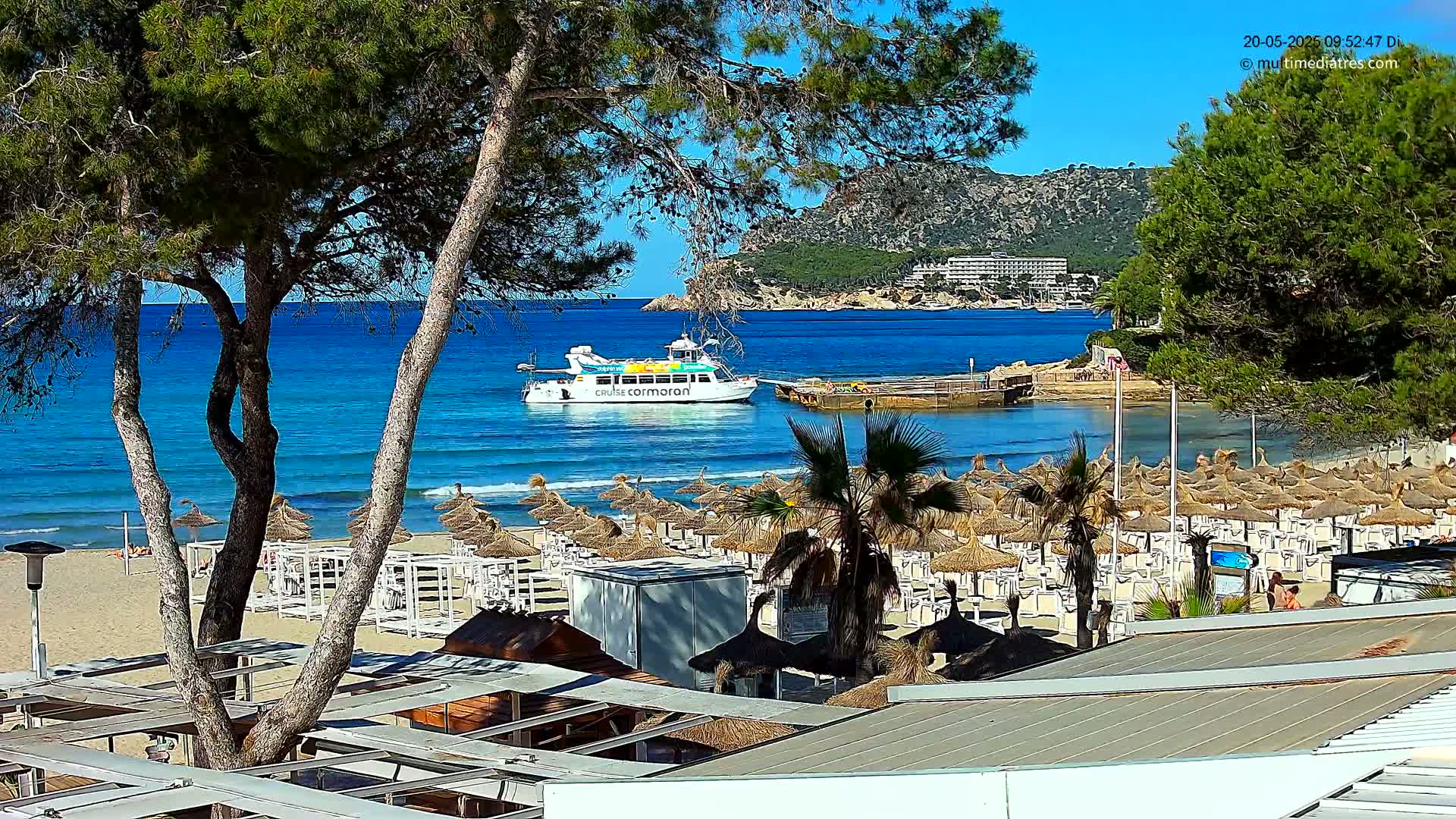 A sandy beach with numerous thatched umbrellas and a boat on a calm, blue ocean under a sunny sky, backed by a hilly landscape with buildings.