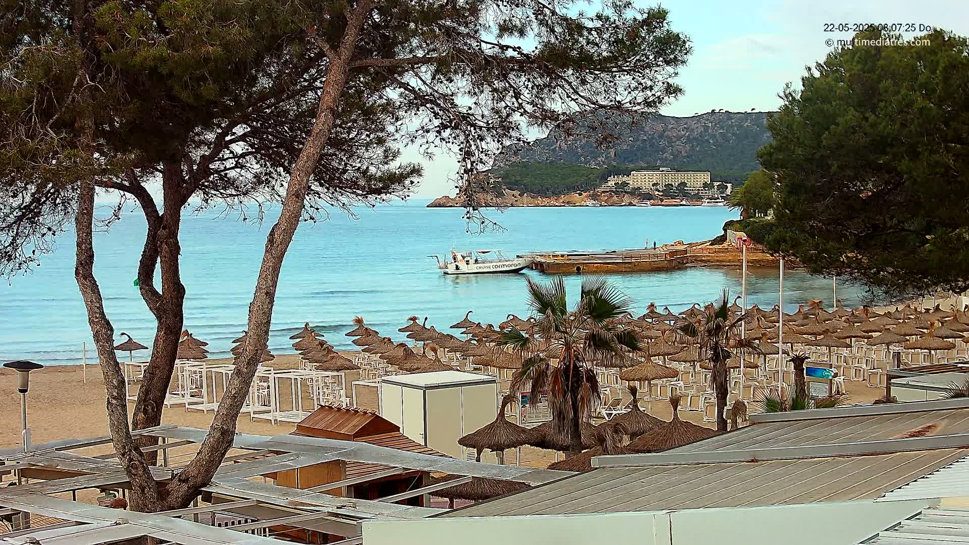 A sandy beach with numerous thatched umbrellas and lounge chairs is situated by a calm, light-blue ocean under a partly cloudy sky; a small boat is visible on the water.