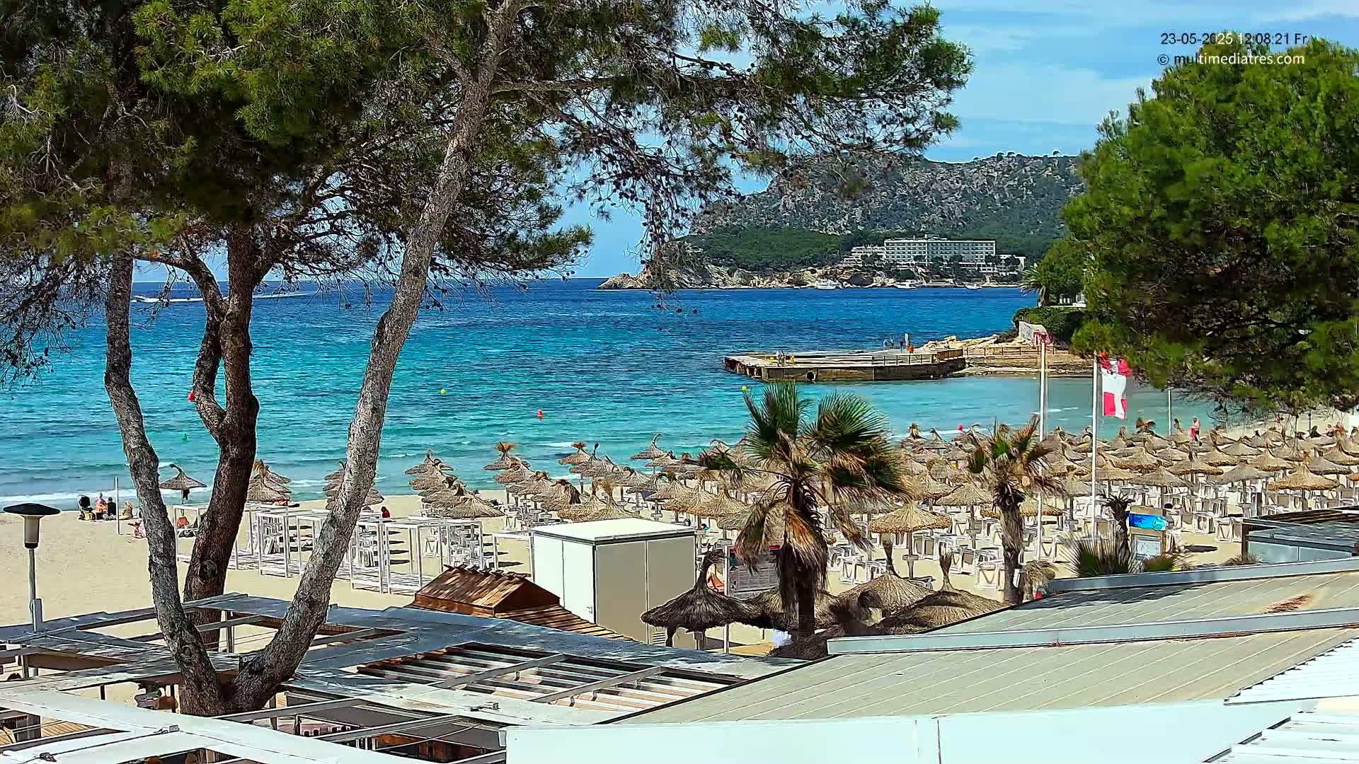 A sandy beach with numerous thatched umbrellas, palm trees, and a clear turquoise ocean under a sunny sky, backed by a rocky hillside.