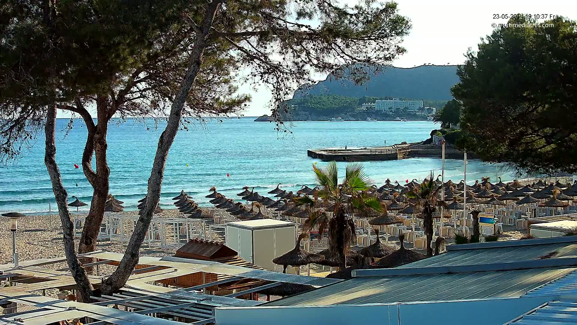A sandy beach with numerous thatched umbrellas and lounge chairs is situated beside a calm, turquoise ocean under a mostly sunny sky, with a rocky hillside visible in the background.