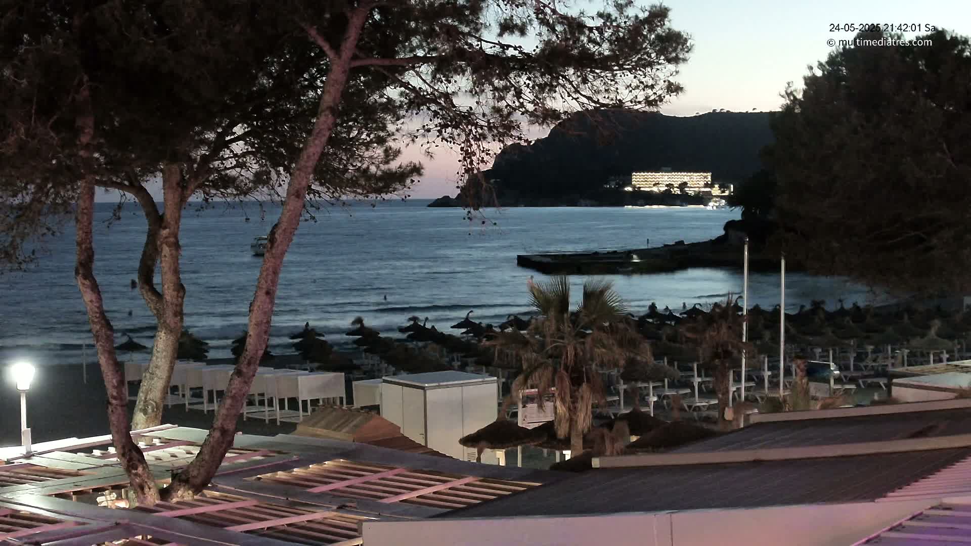 A nighttime beach scene shows a calm sea under a dusky sky, with a hotel visible in the distance, beach umbrellas and lounge chairs on the sand, and trees in the foreground.
