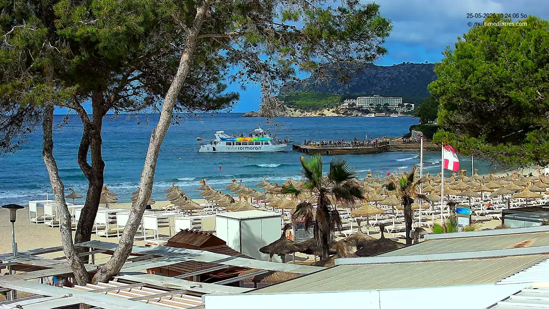 A sandy beach with numerous thatched umbrellas and lounge chairs is situated near calm, blue ocean water, with a passenger boat visible offshore under a partly cloudy sky.
