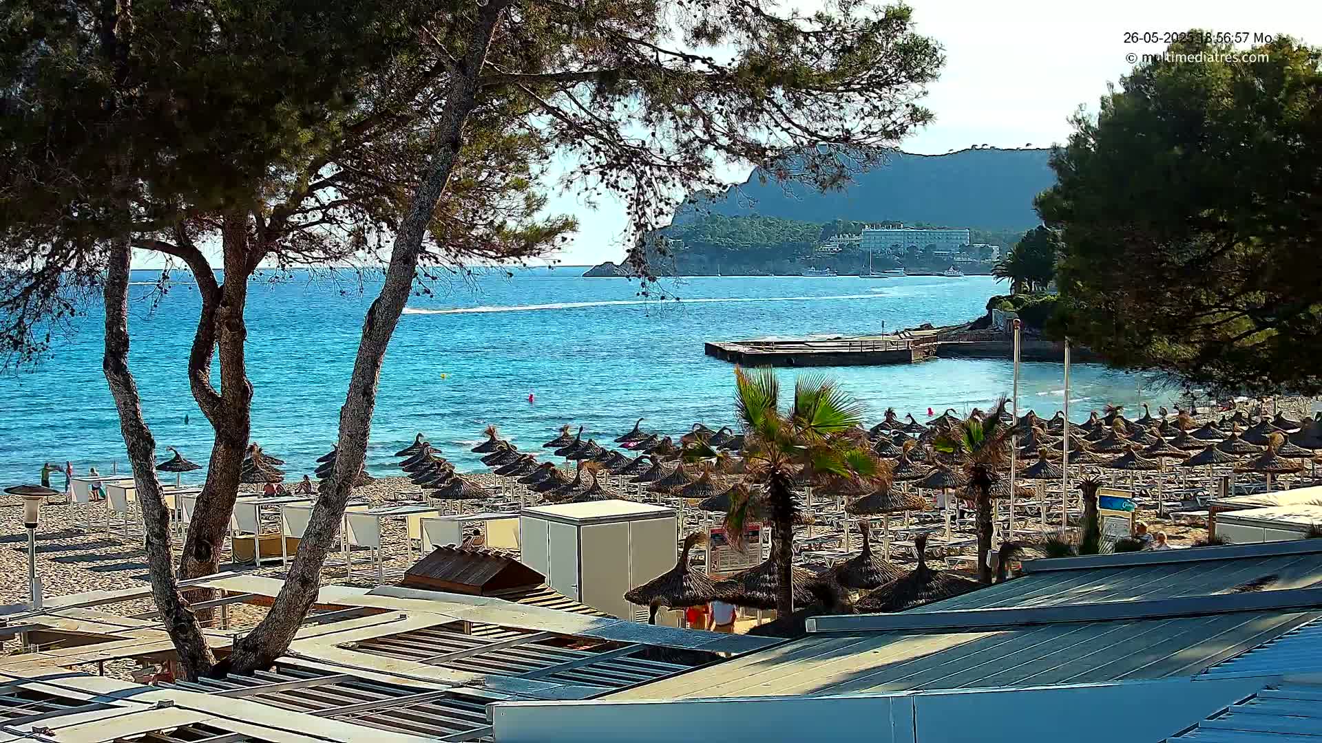 A sandy beach with numerous thatched umbrellas and palm trees overlooks a calm, turquoise ocean under a sunny sky, backed by a distant mountain range.
