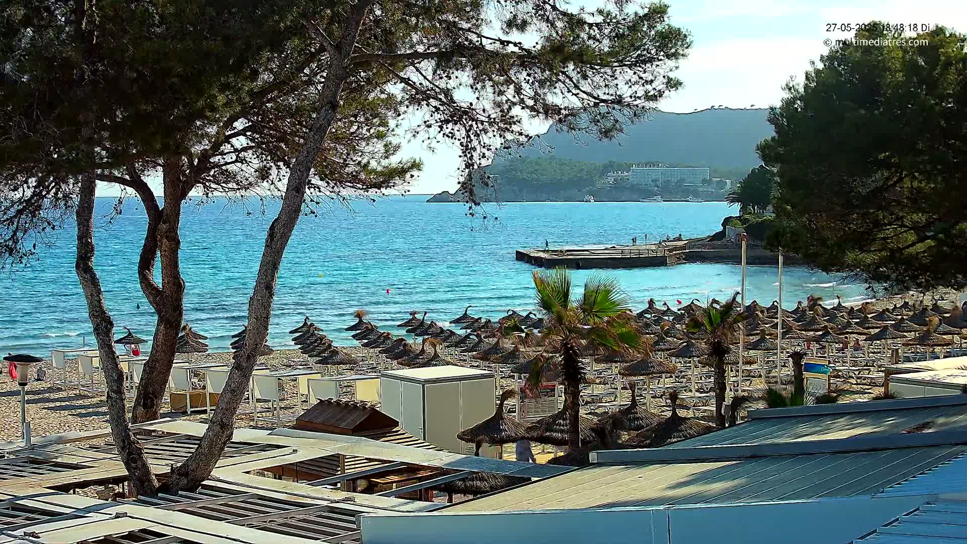 A sandy beach with numerous thatched umbrellas and lounge chairs is situated beside a calm, turquoise ocean under a sunny sky, backed by a rocky hillside.