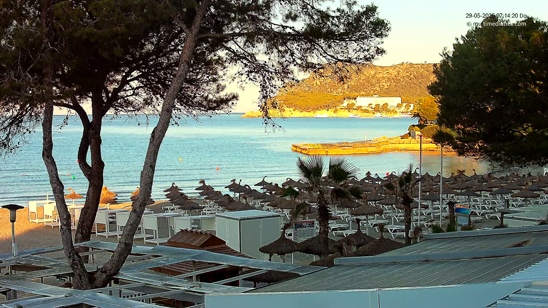 A beach with numerous thatched umbrellas and lounge chairs is situated next to a calm, clear ocean under a partly sunny sky; a hilly coastline is visible in the background.