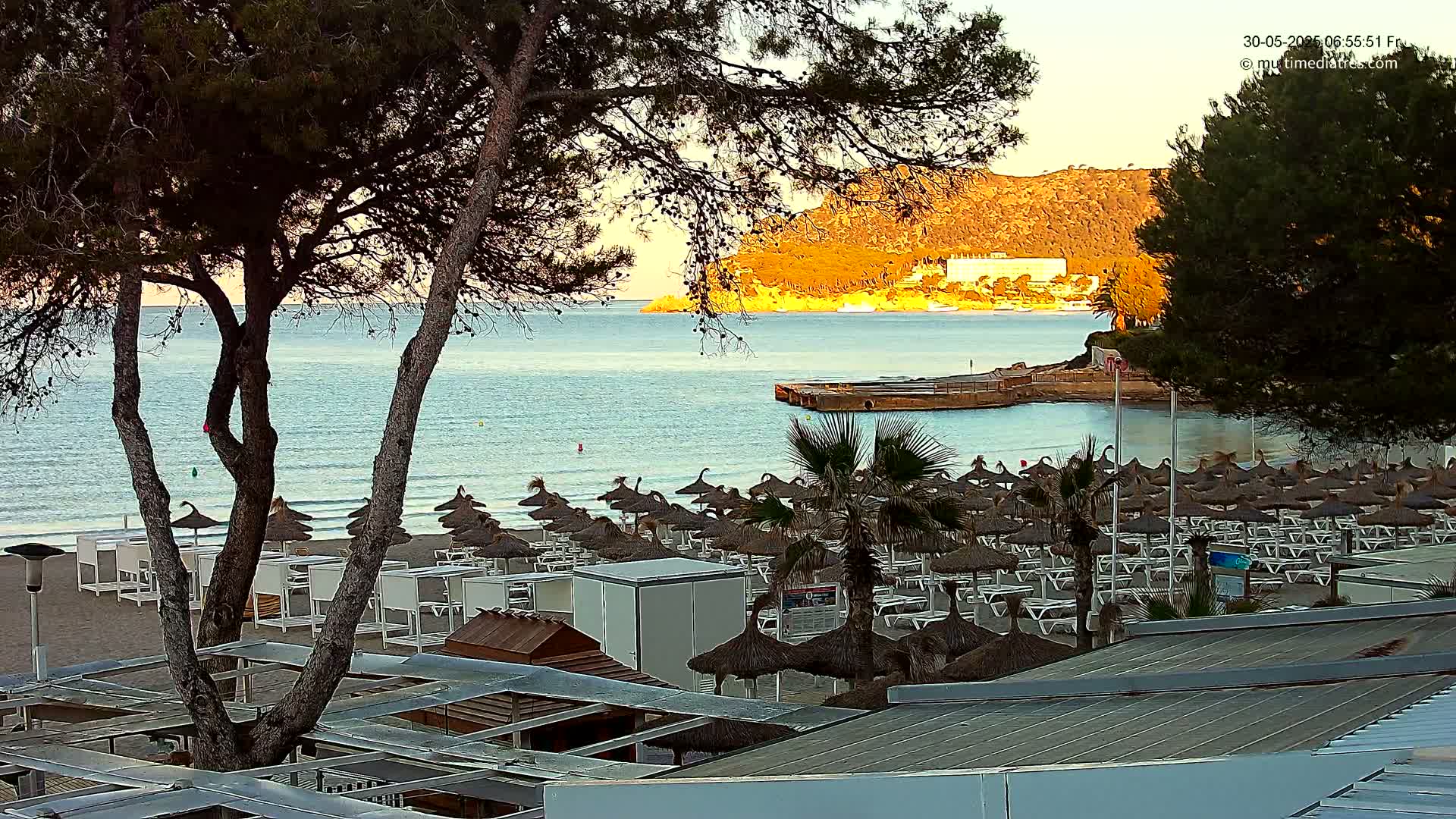 A sandy beach with numerous umbrellas and lounge chairs is situated beside a calm sea, with a hill in the background under a clear, sunrise sky.