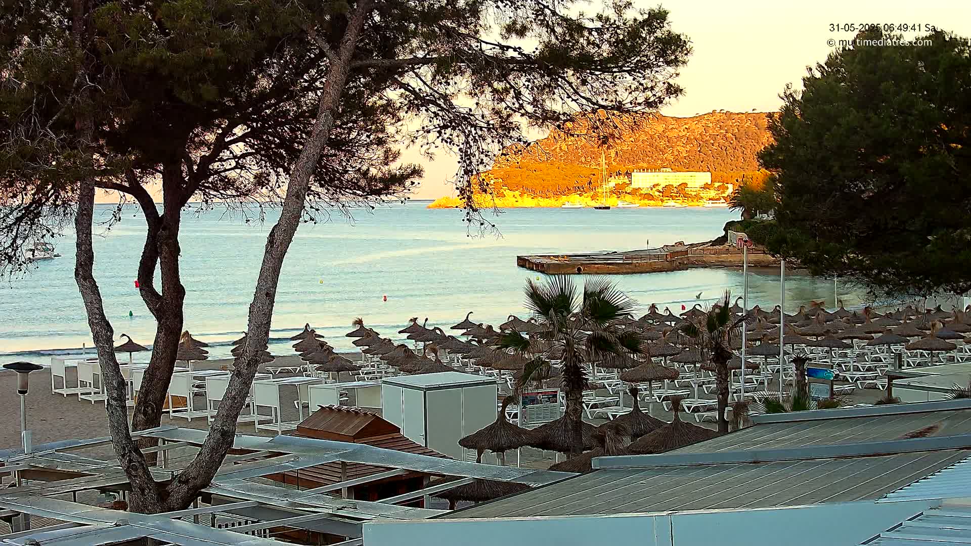 A sandy beach with numerous thatched umbrellas and lounge chairs is situated near calm, pale-blue water, under a tranquil, sunrise sky, with a hill in the background.