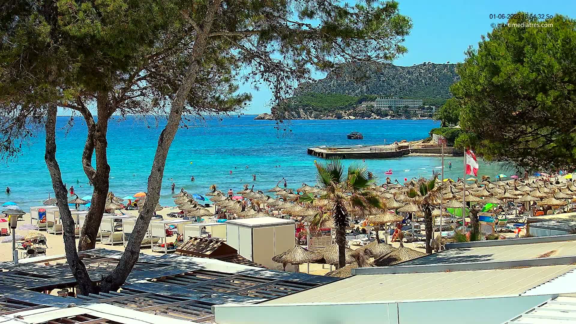 A sunny beach scene shows a crowded sandy shore with numerous straw umbrellas, a calm turquoise sea, and green hills in the background.