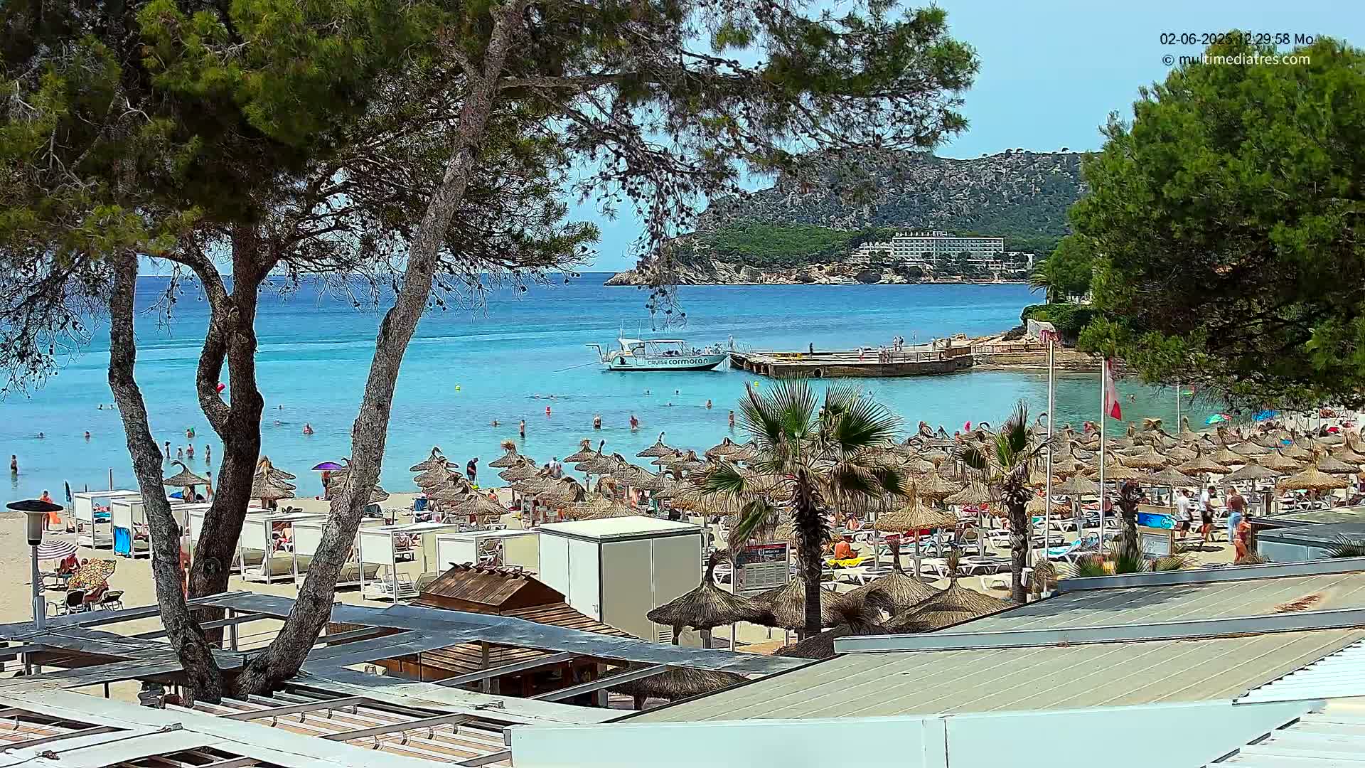A sunny day at a crowded beach with turquoise water, numerous thatched umbrellas, and a boat in the bay.