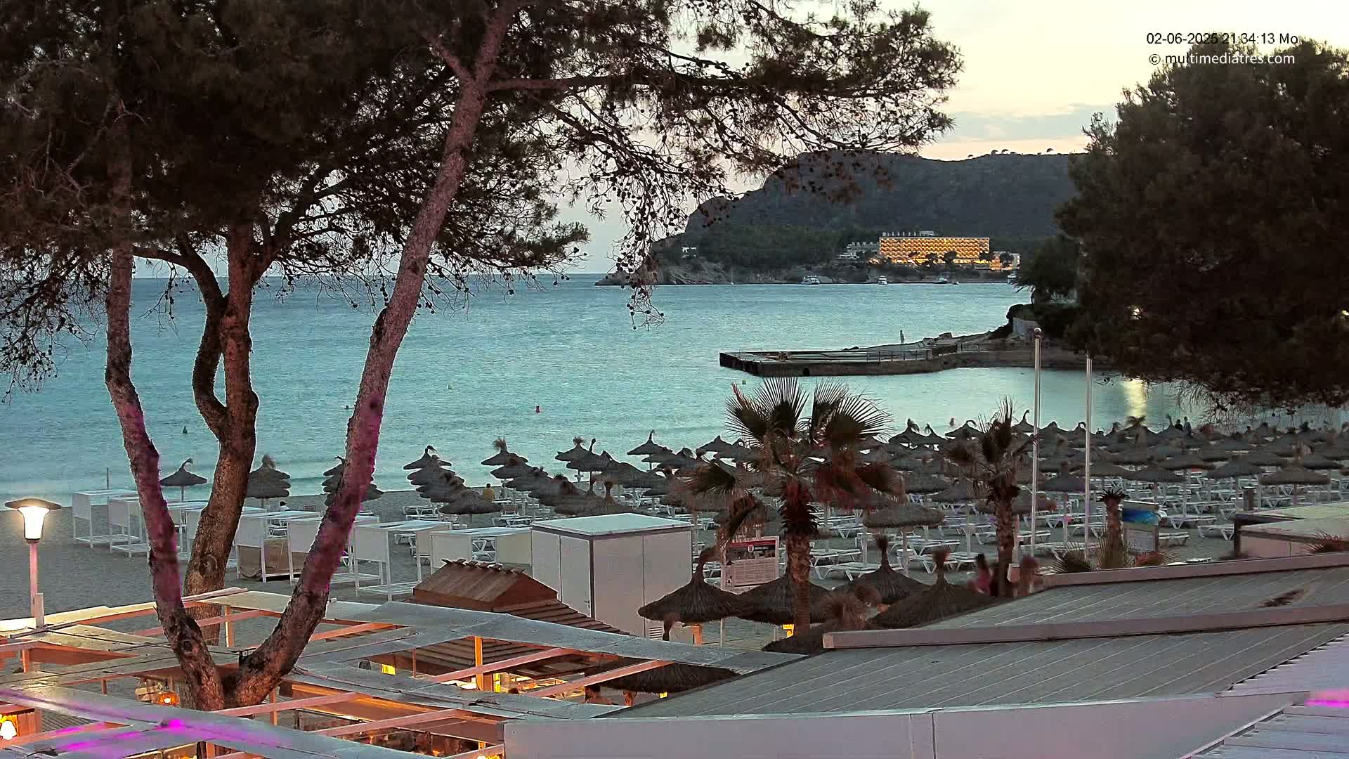 A beach at dusk, with numerous umbrellas and lounge chairs, overlooks a calm sea and a distant building on a hillside.
