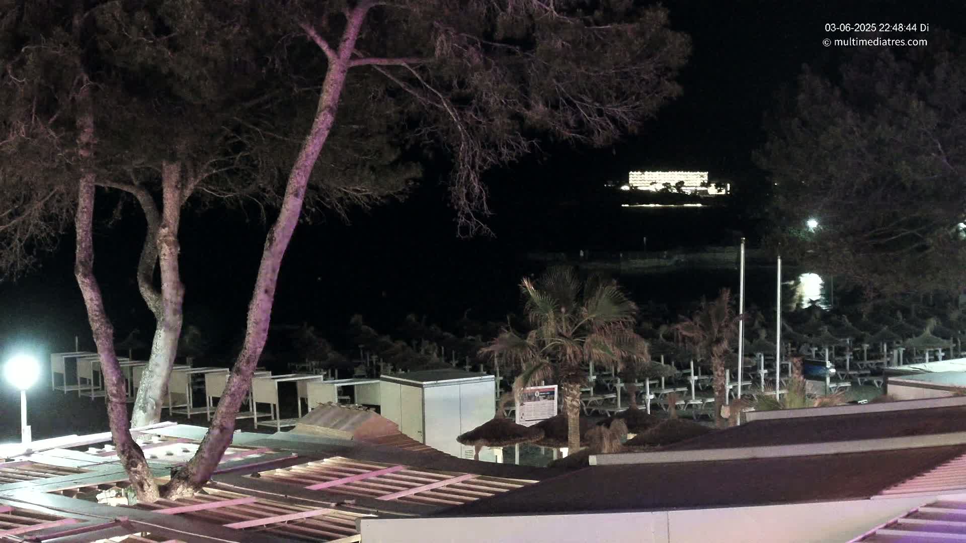 A nighttime view of a beach at night, with dark trees, closed beach umbrellas, and a brightly lit building in the distance.