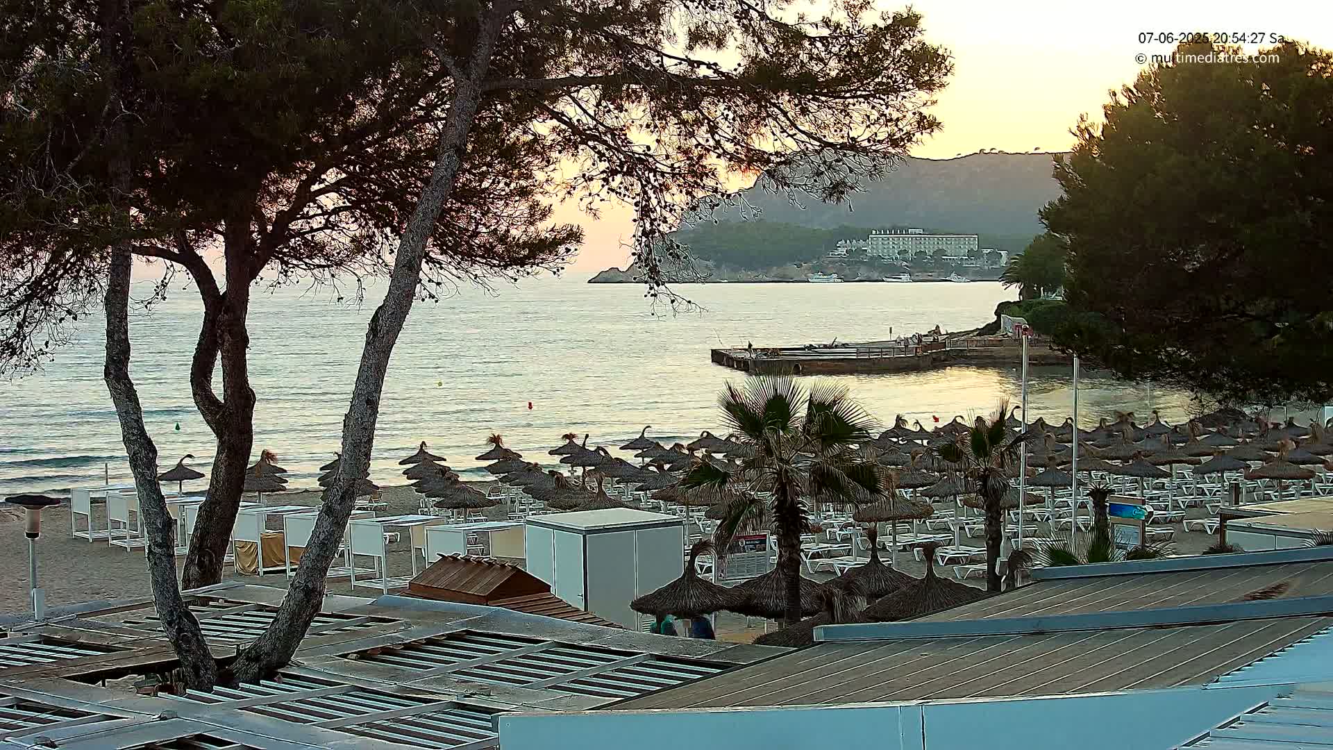 A calm ocean at sunset with a sandy beach lined with numerous thatched umbrellas and lounge chairs, viewed from behind trees.