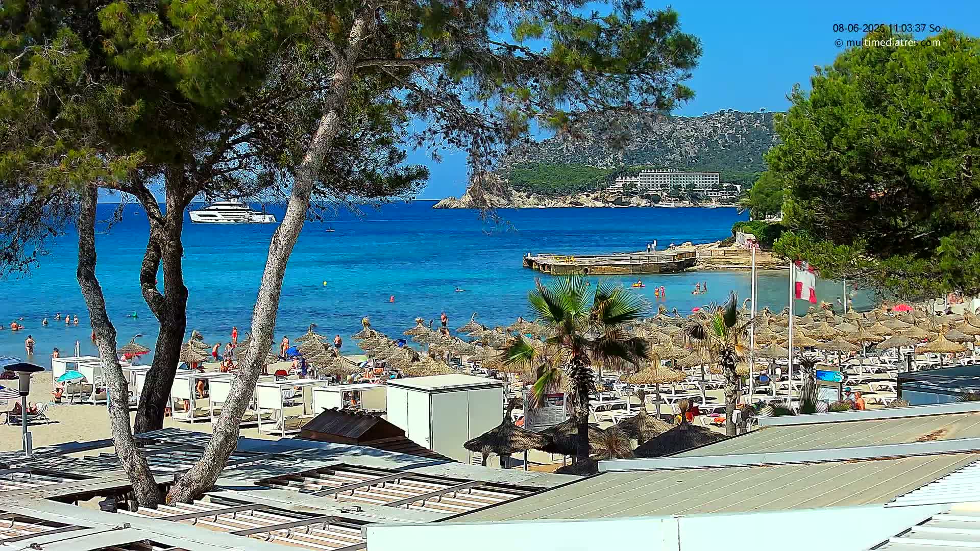 A sunny beach scene shows a sandy shore lined with thatched umbrellas and lounge chairs, a calm turquoise sea with swimmers, a distant yacht, and a hillside with buildings in the background.