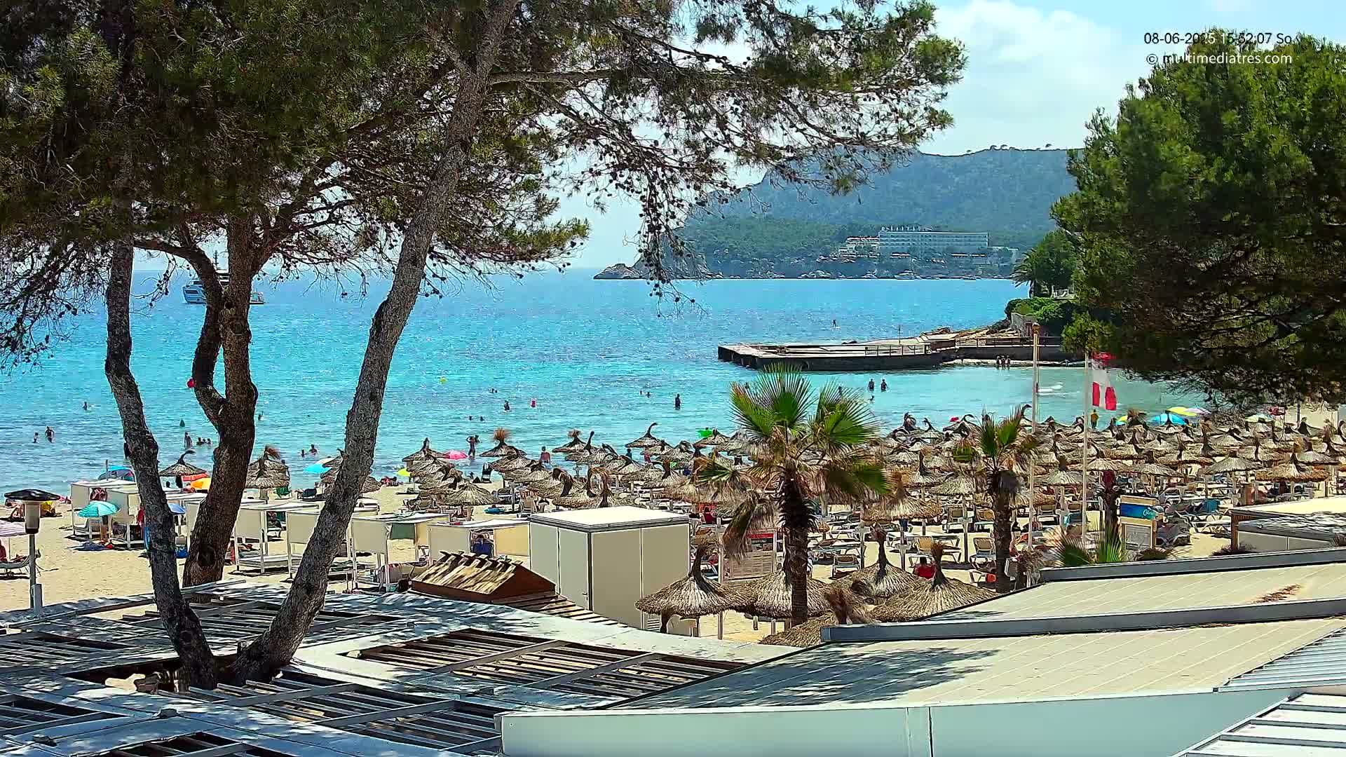 A sunny beach scene shows a sandy shore lined with numerous thatched umbrellas, palm trees, and people swimming in clear, turquoise water, with a rocky hillside and distant buildings visible beyond.