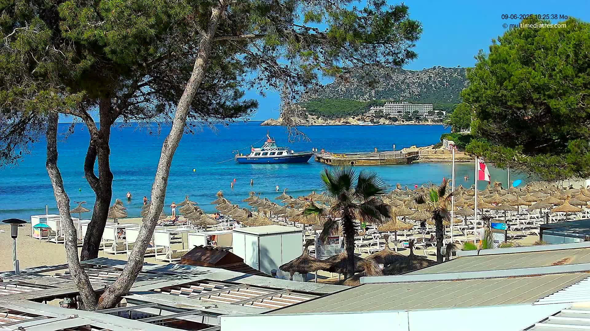 A sandy beach with numerous thatched umbrellas and lounge chairs is viewed from above, showcasing a calm, turquoise ocean with a boat and distant hills under a sunny sky.