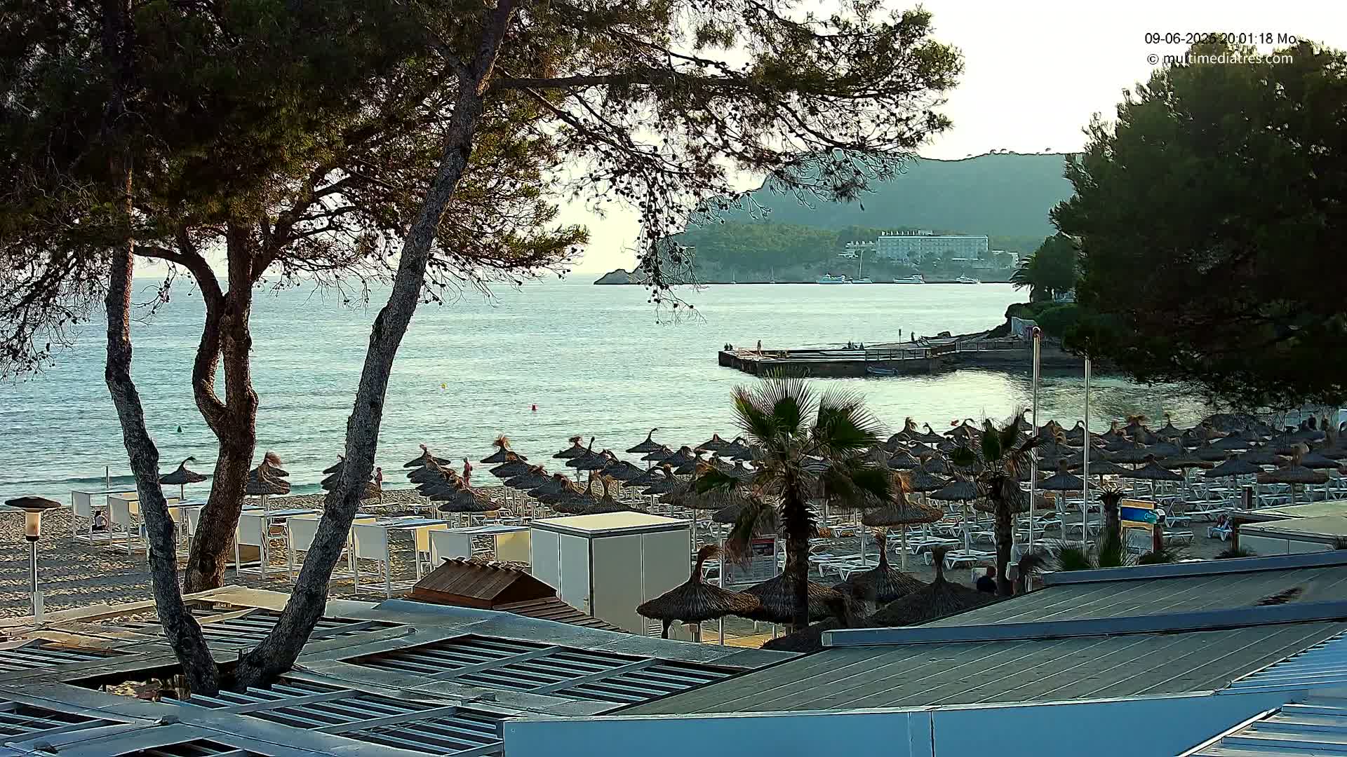 A sandy beach with numerous thatched umbrellas and lounge chairs sits under a calm, clear evening sky next to a calm sea with a small pier and distant hills.