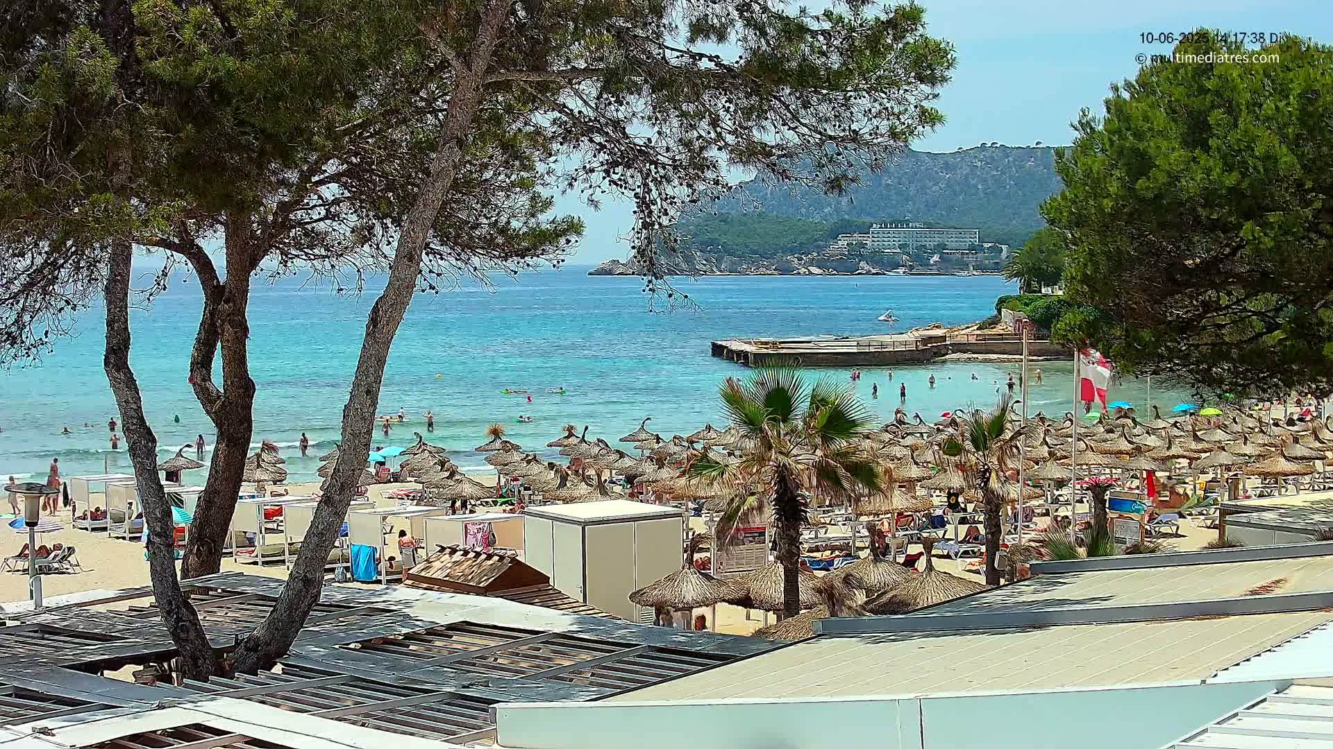 A sunny beach scene shows a calm, turquoise sea, a sandy shore dotted with numerous thatched umbrellas and lounge chairs, and a backdrop of green trees and distant hills.