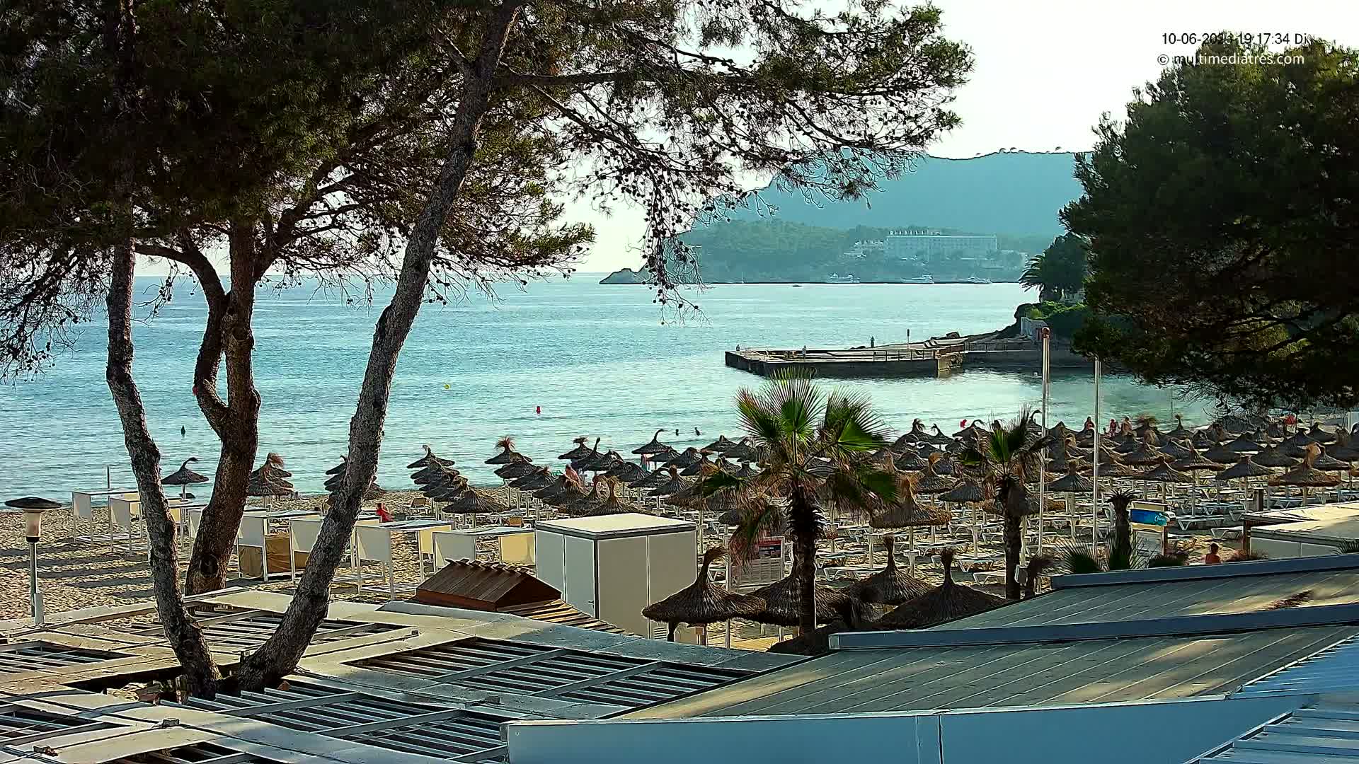 A sandy beach with numerous thatched umbrellas and lounge chairs is seen from above, overlooking a calm, light blue sea, backed by green hills and trees under a clear sky.