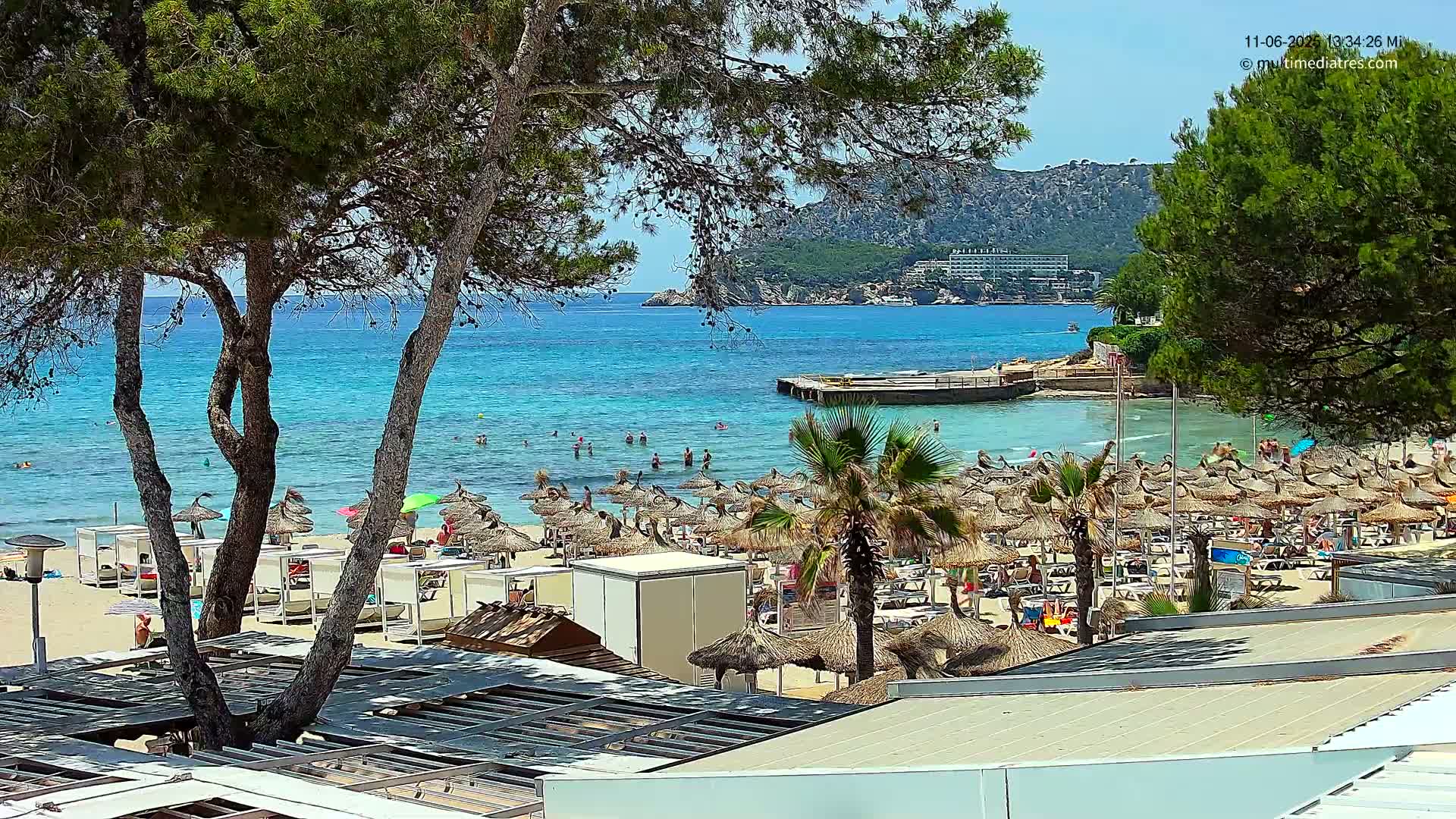 A sandy beach with numerous thatched umbrellas and sunbathers is situated next to clear turquoise water, under a sunny sky, with trees and hills visible in the background.