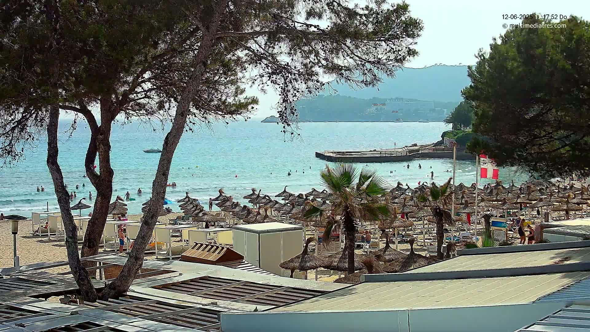 A sandy beach with numerous thatched umbrellas, palm trees, and bathers in calm, clear turquoise water under a mostly sunny sky, overlooked by trees and a distant mountain.
