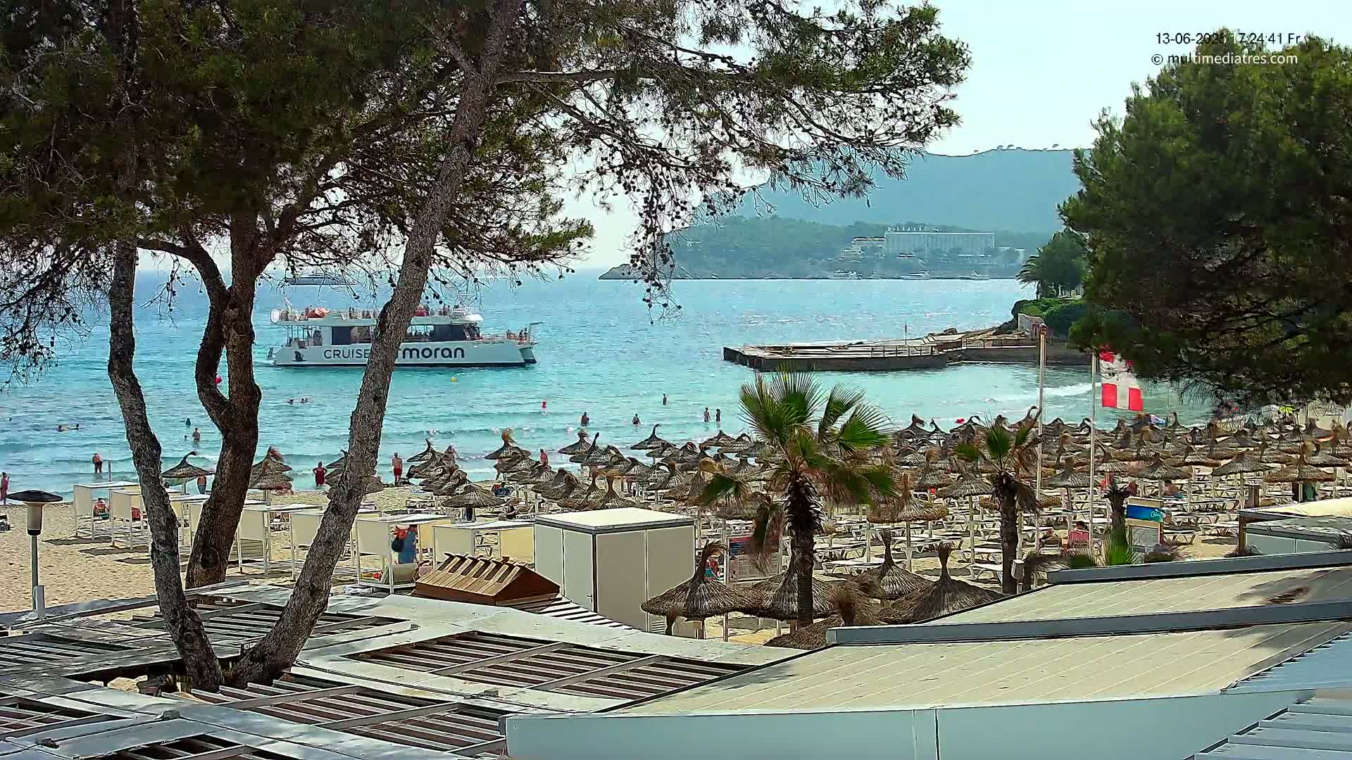 A sunny beach scene shows a boat on calm blue water, numerous beach umbrellas and people on the sand, and palm trees and pine trees in the foreground.