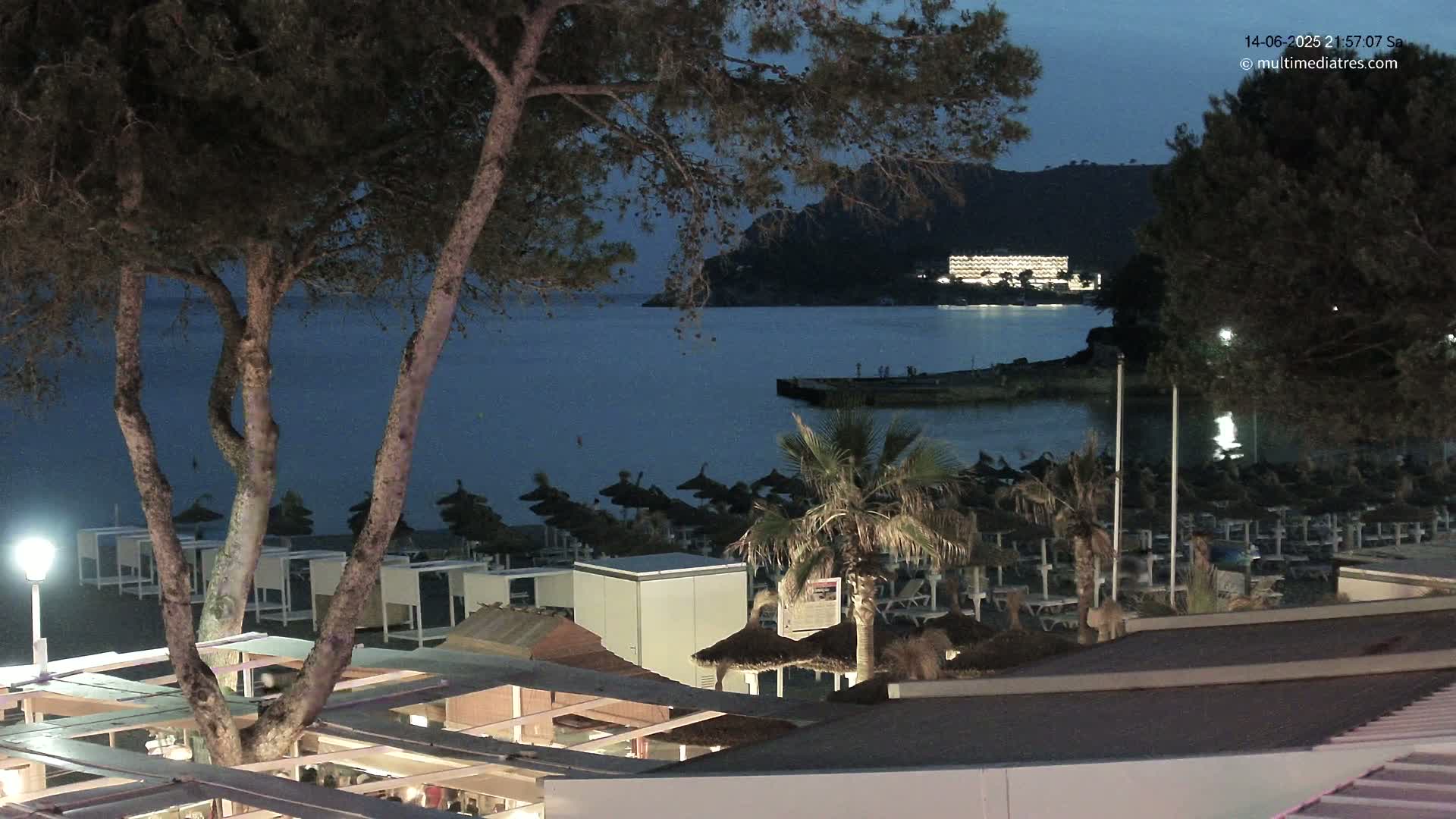 A sunny beach scene shows a calm, turquoise ocean with many people swimming and relaxing under numerous thatched umbrellas, a boat docked nearby, and pine trees framing the view.