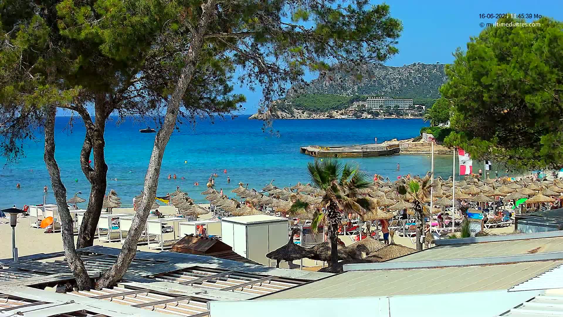 A sunny beach scene shows a sandy shore lined with numerous straw umbrellas and beachgoers, a calm turquoise sea with swimmers, and a rocky headland in the background.