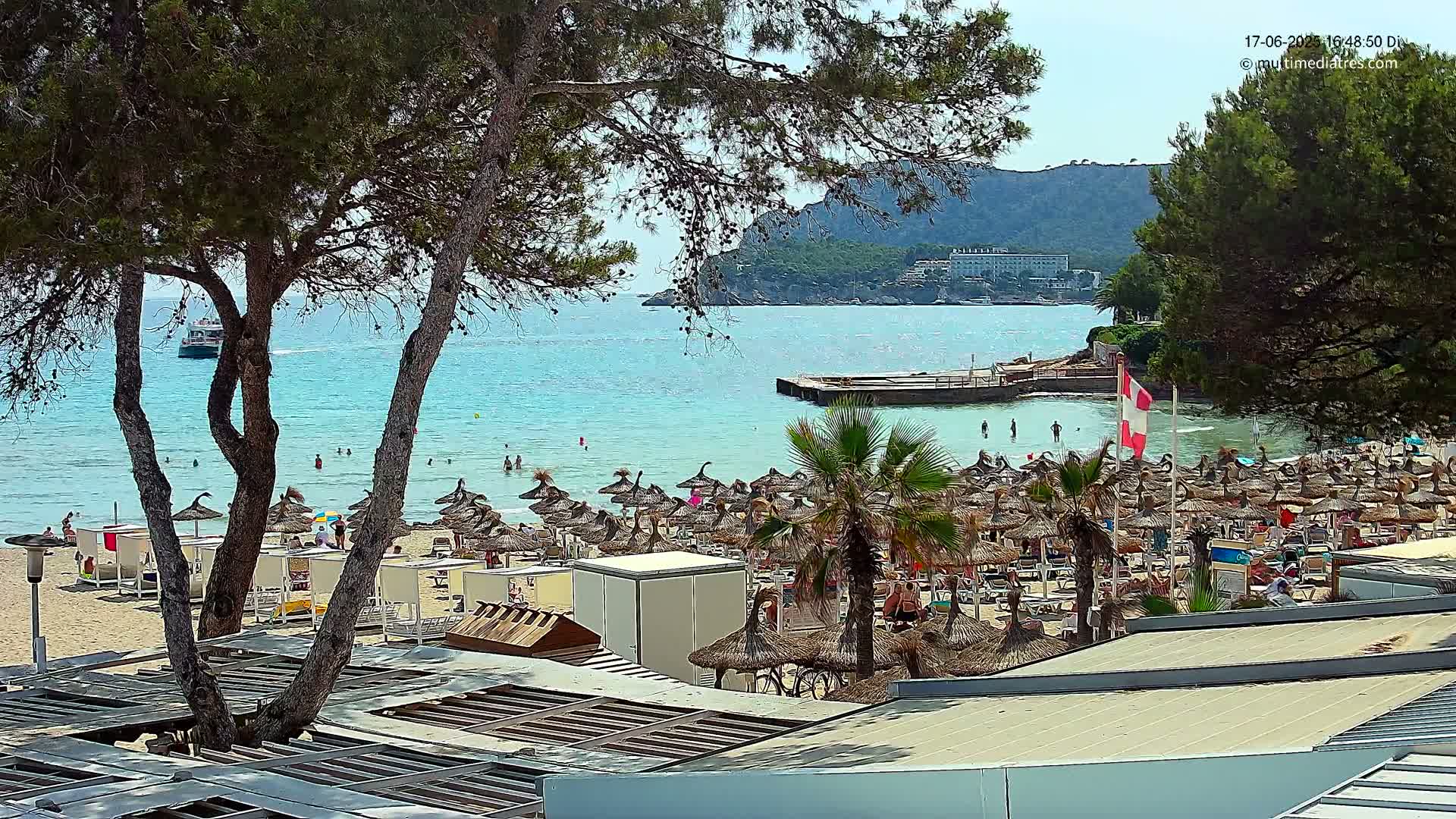 A sandy beach with numerous thatched umbrellas, palm trees, and people swimming in clear turquoise water under a sunny sky, backed by green hills and a distant boat.