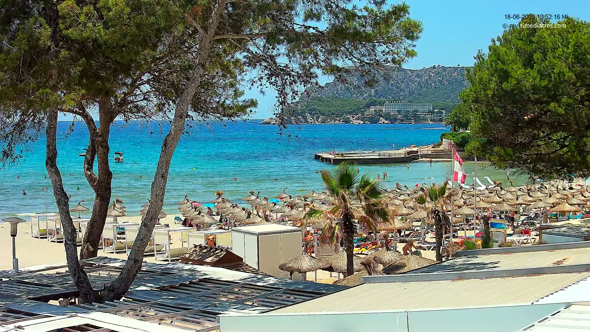 A sunny beach scene shows a sandy shore covered in numerous thatched umbrellas, palm trees, and bathers, all situated next to a calm, turquoise sea with a rocky coastline and distant hills in the background.