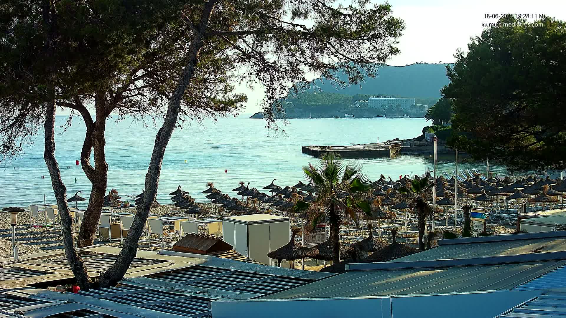 A beach with many thatched umbrellas and lounge chairs sits beside a calm, clear body of water under a sunny sky, with green hills visible in the background.
