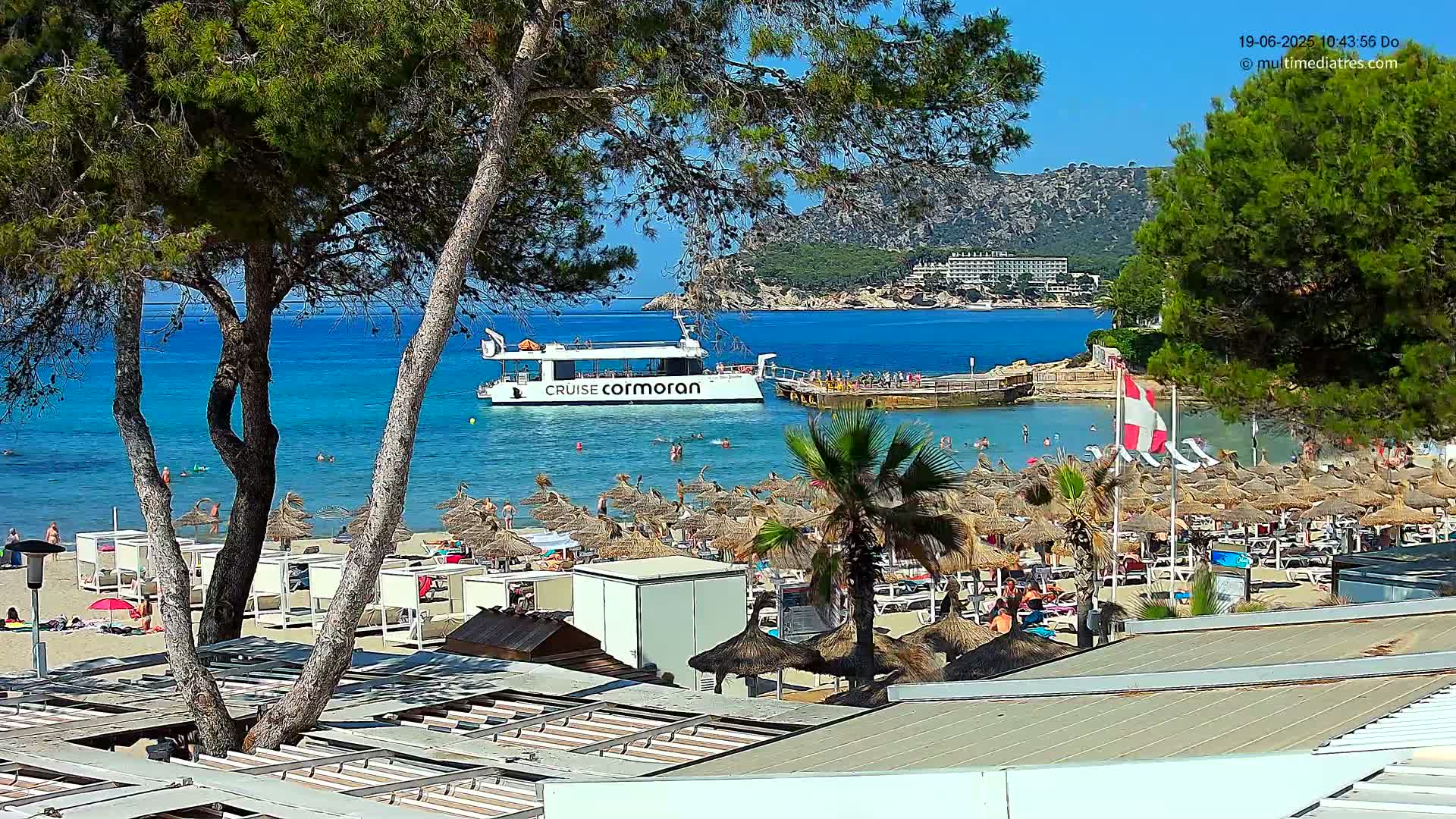 A sunny beach scene shows numerous thatched umbrellas, beachgoers, and a boat named "Cruise Cormoran" in calm, clear turquoise water, with pine trees and a distant hillside visible.
