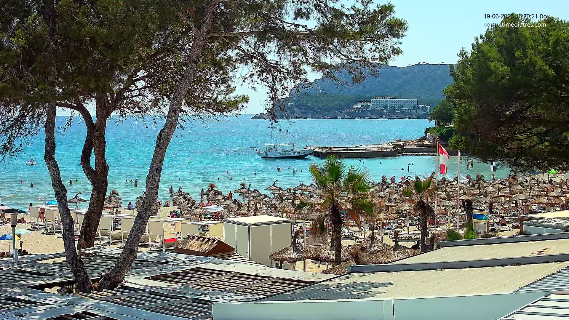 A sunny day at a crowded beach with turquoise water, numerous beach umbrellas, and a boat in the bay, all viewed from behind trees.