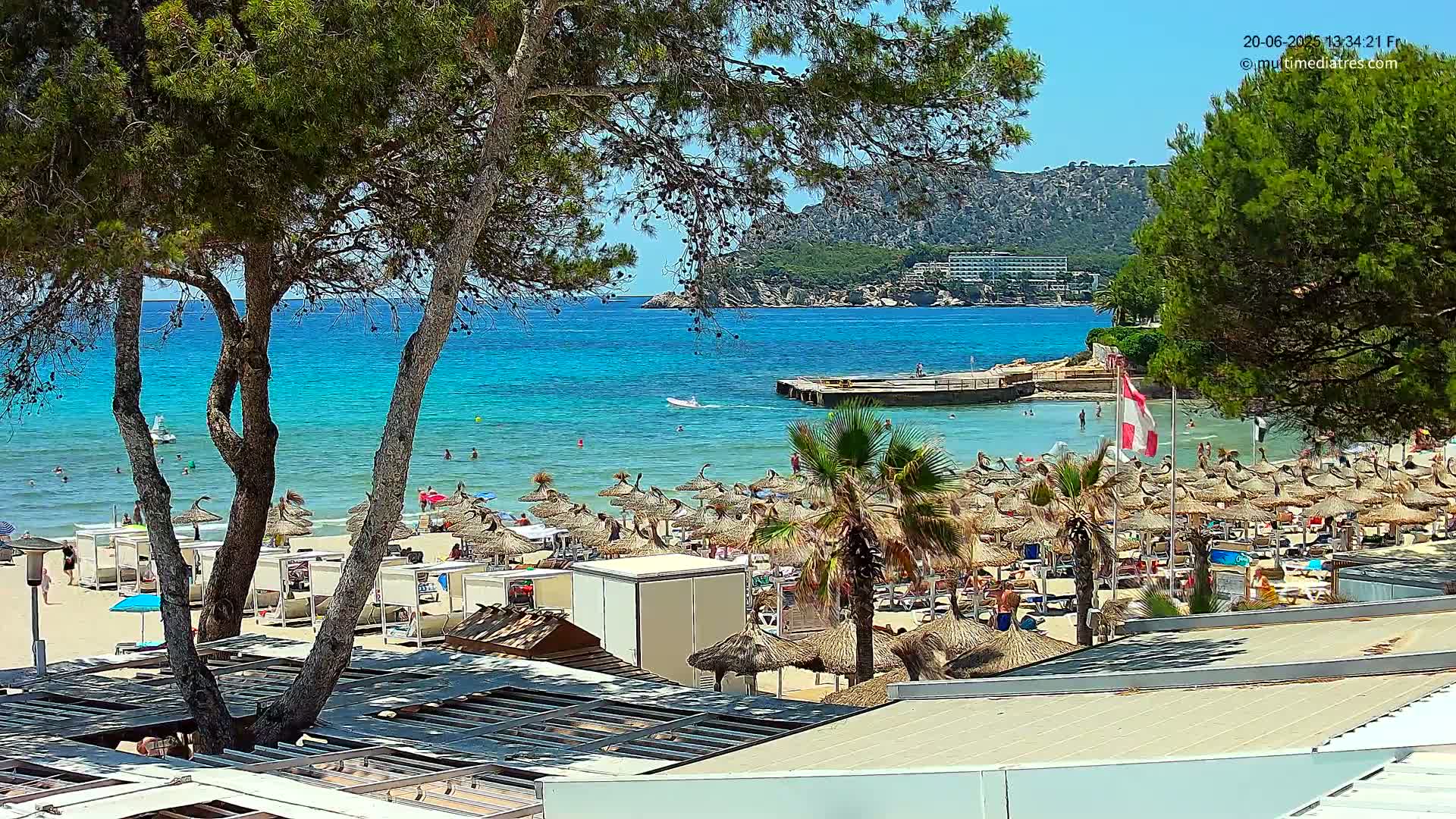 A sunny beach scene shows a sandy shore lined with numerous thatched umbrellas, palm trees, and people enjoying the clear, turquoise water under a bright sky.