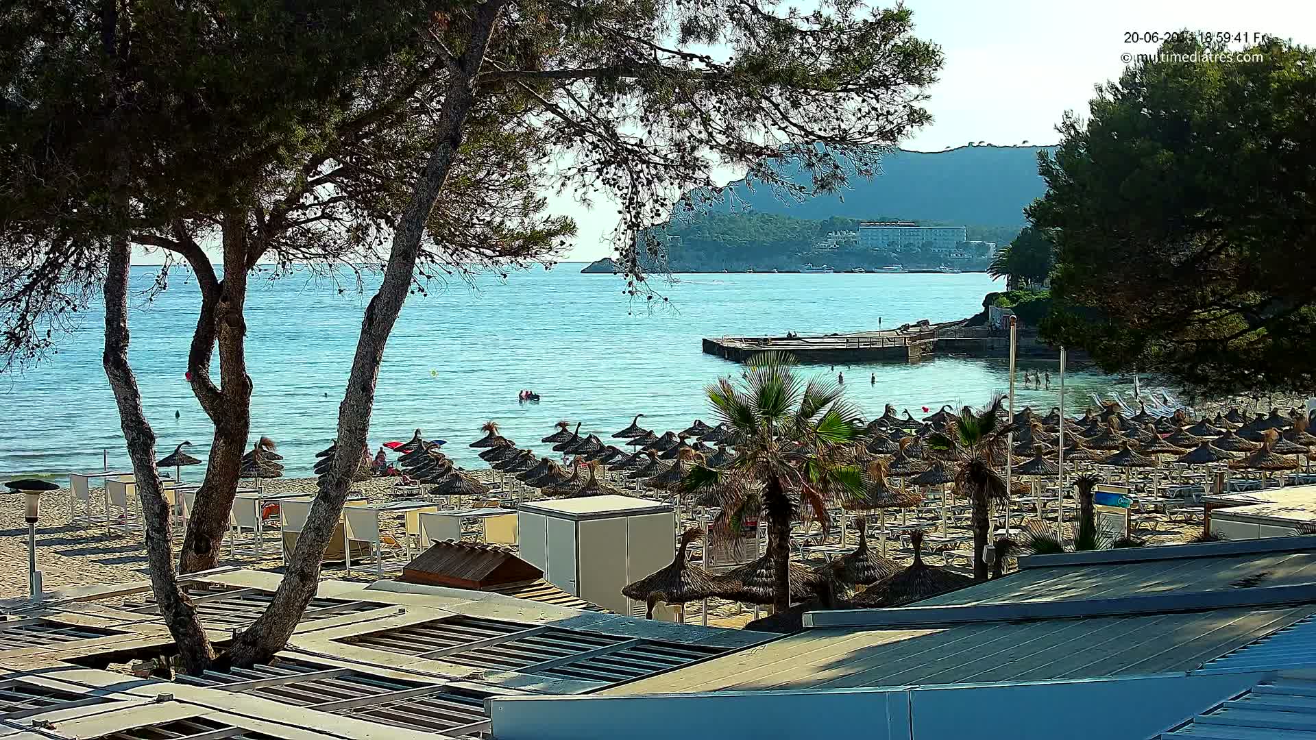A sandy beach with numerous thatched umbrellas and lounge chairs overlooks a calm, turquoise sea under a sunny sky, backed by a distant mountainous landscape.