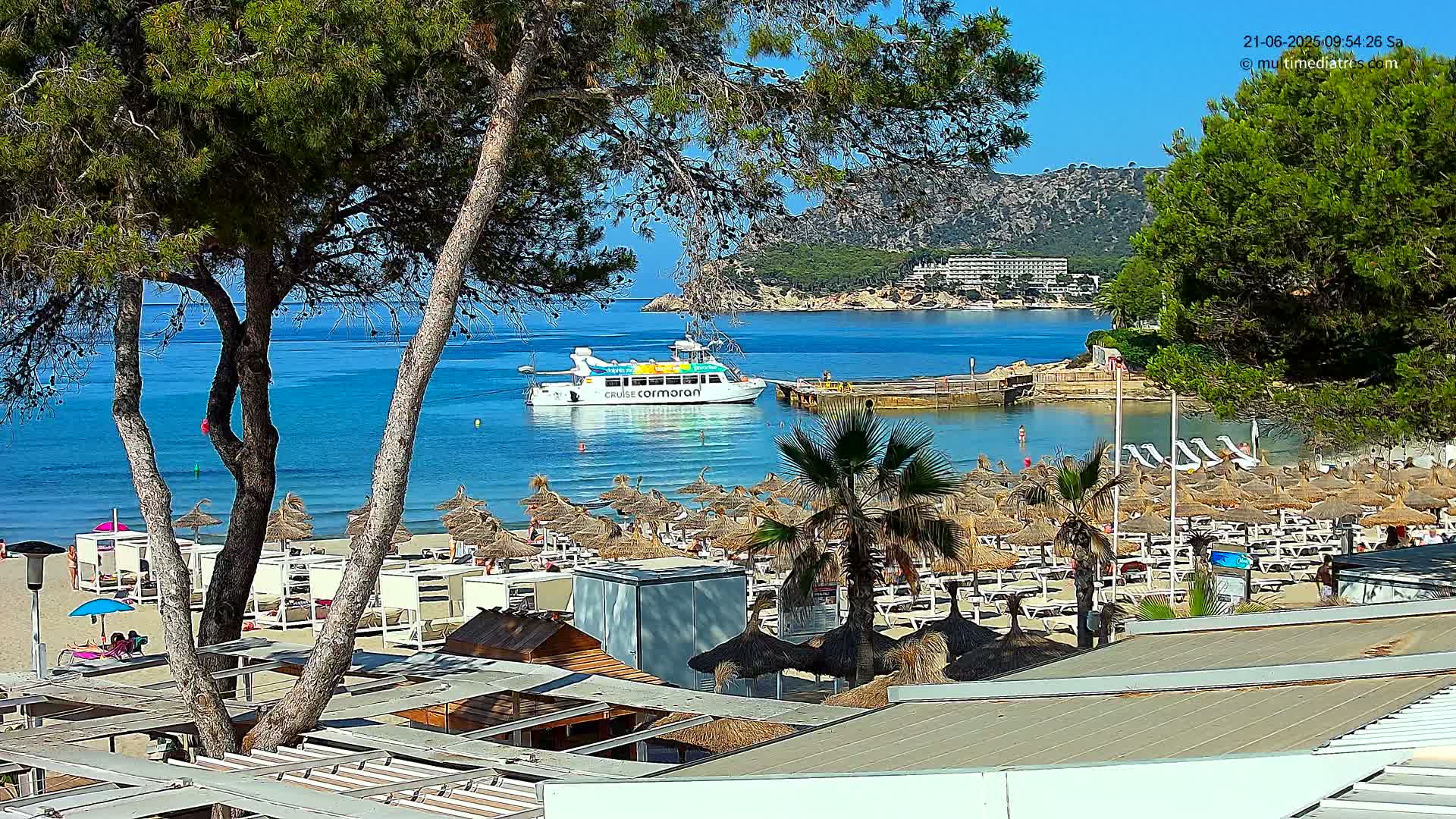 A sandy beach with numerous thatched umbrellas and lounge chairs, partially obscured by pine trees, overlooks a calm, blue bay where a passenger boat is moored near a small dock and a hillside with buildings in the distance under a clear blue sky.