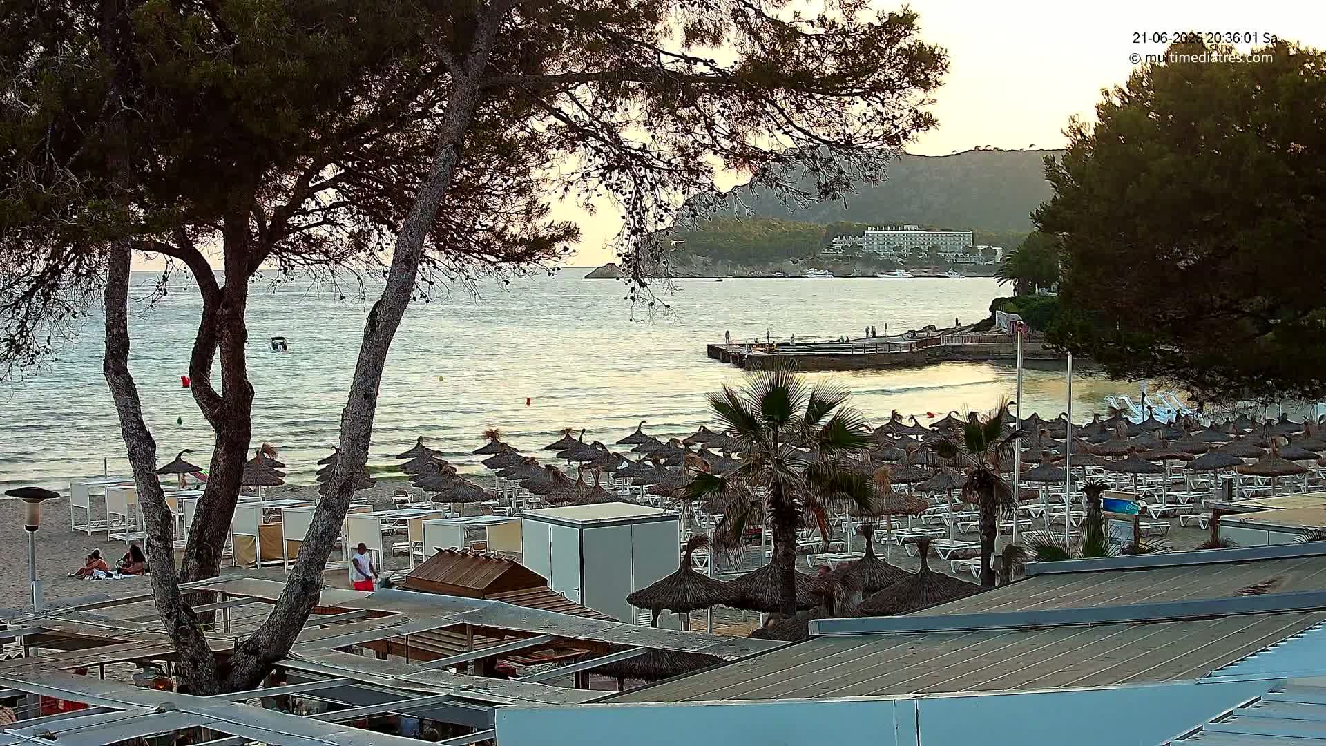 A sandy beach with numerous thatched umbrellas and lounge chairs is situated beside calm ocean water under a partly cloudy evening sky, with a distant hotel visible across the bay.