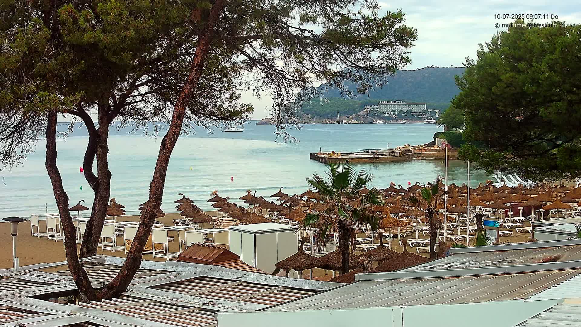 A sandy beach with numerous thatched umbrellas and lounge chairs overlooks a calm sea under a cloudy sky, with trees in the foreground and a distant hotel visible on a hillside.