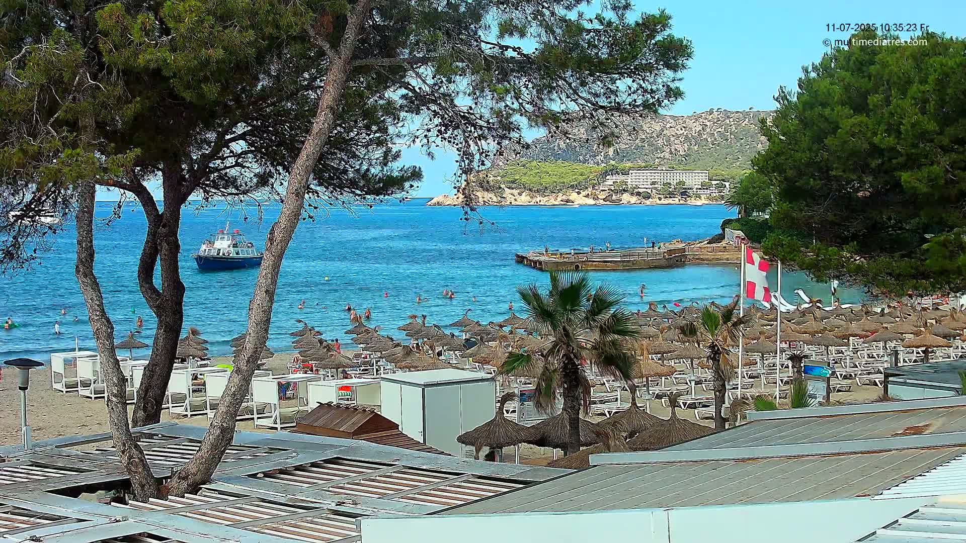 A sunny beach scene shows a calm, blue ocean with a boat, numerous beach umbrellas and lounge chairs, and palm trees, all nestled beside a rocky hillside.
