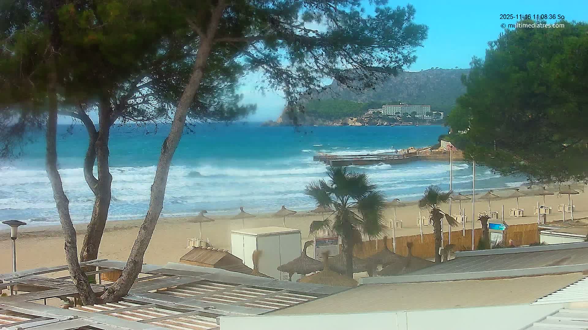 The image displays a sunny day with a clear blue sky over a sandy beach featuring choppy turquoise waves, framed by green trees and scattered straw umbrellas, with a pier and a distant hotel visible across the bay.