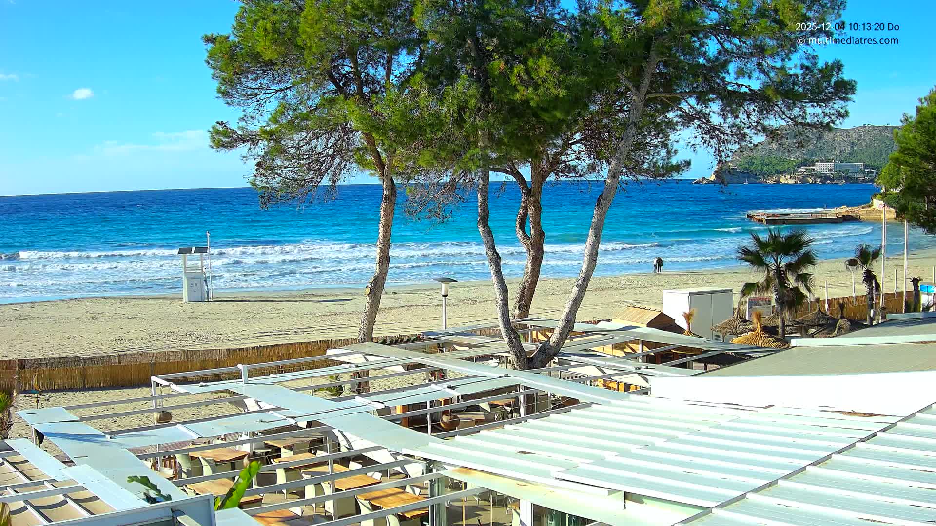 On a clear and sunny day, an outdoor restaurant patio overlooks a sandy beach where gentle blue waves break, flanked by pine trees, a lifeguard stand, and a few people strolling, with distant cliffs, buildings, and a pier completing the scenic view.