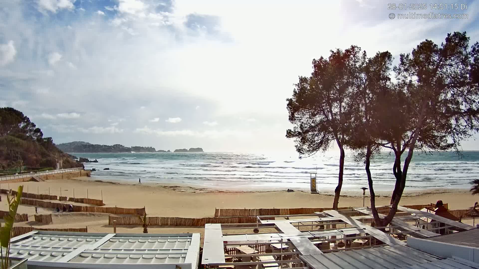 A sandy beach with moderate waves under a partly cloudy sky is viewed from behind a wooden fence and partially-covered outdoor seating area, with several pine trees in the foreground.