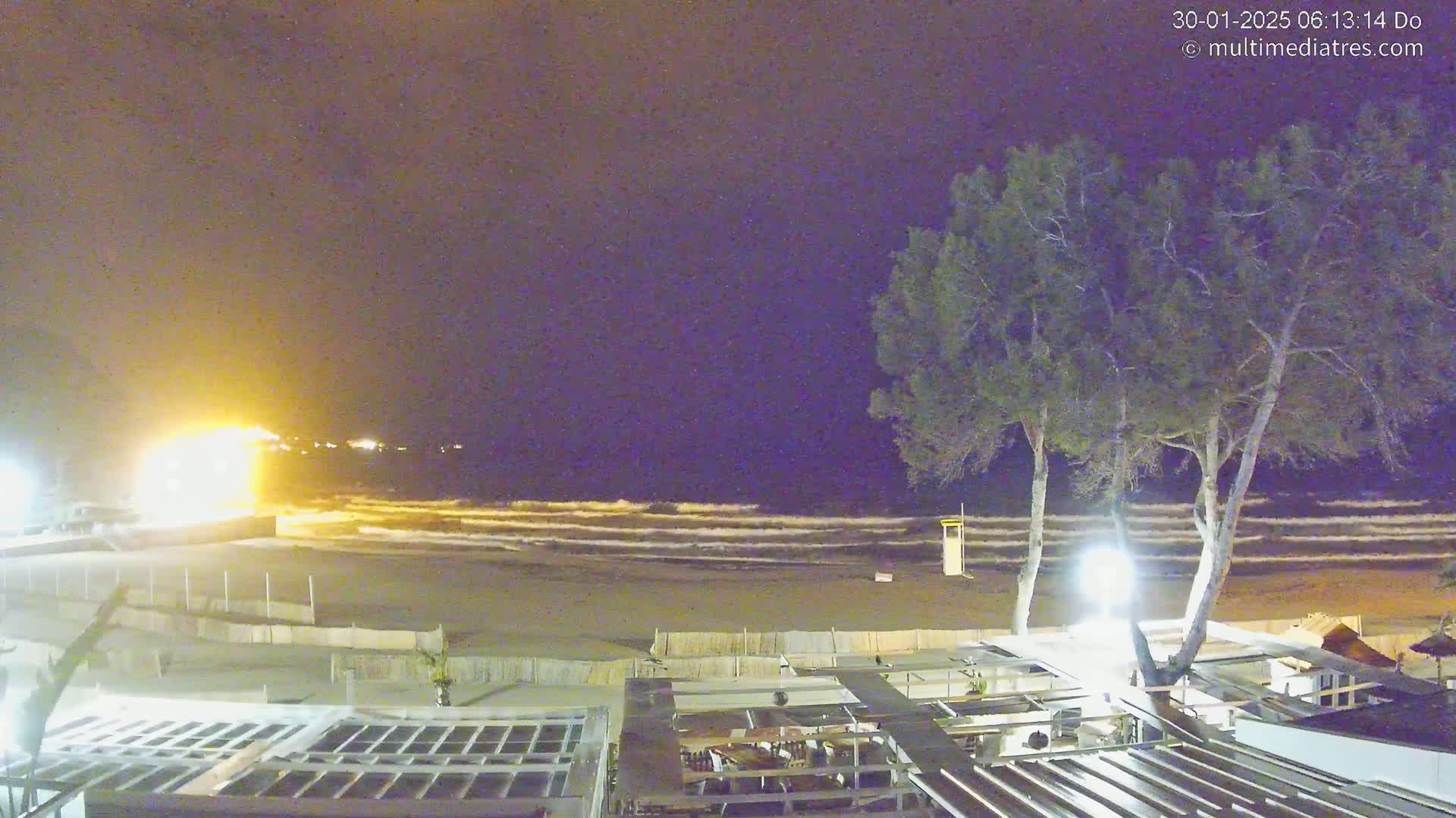 A nighttime beach scene with moderate waves under a dark sky shows a beachside restaurant partially illuminated with lights, and two trees standing between the building and the ocean.